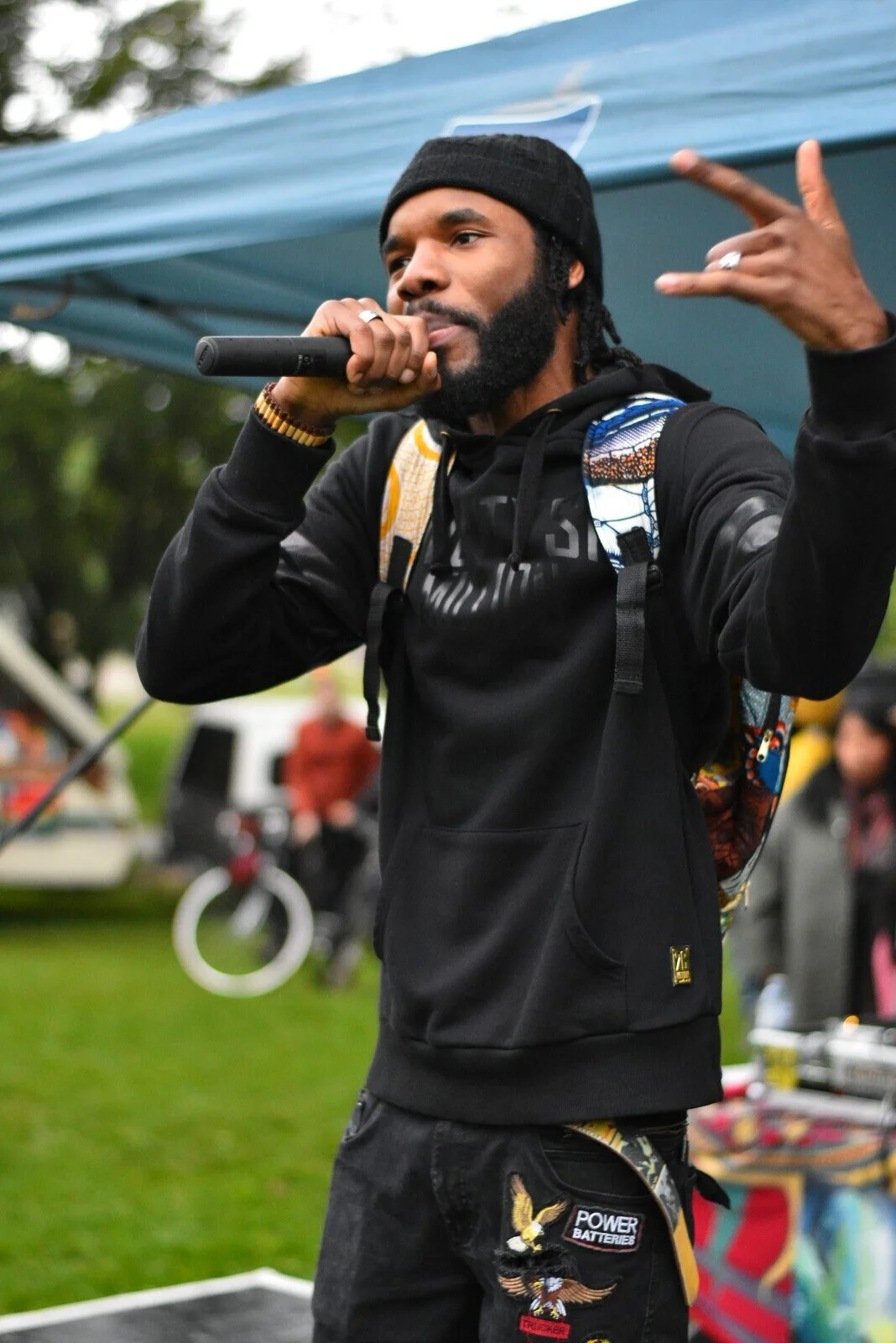 A man wearing a black hoodie and beanie, holding a microphone and gesturing with his other hand, appears to be performing or speaking at an outdoor event.