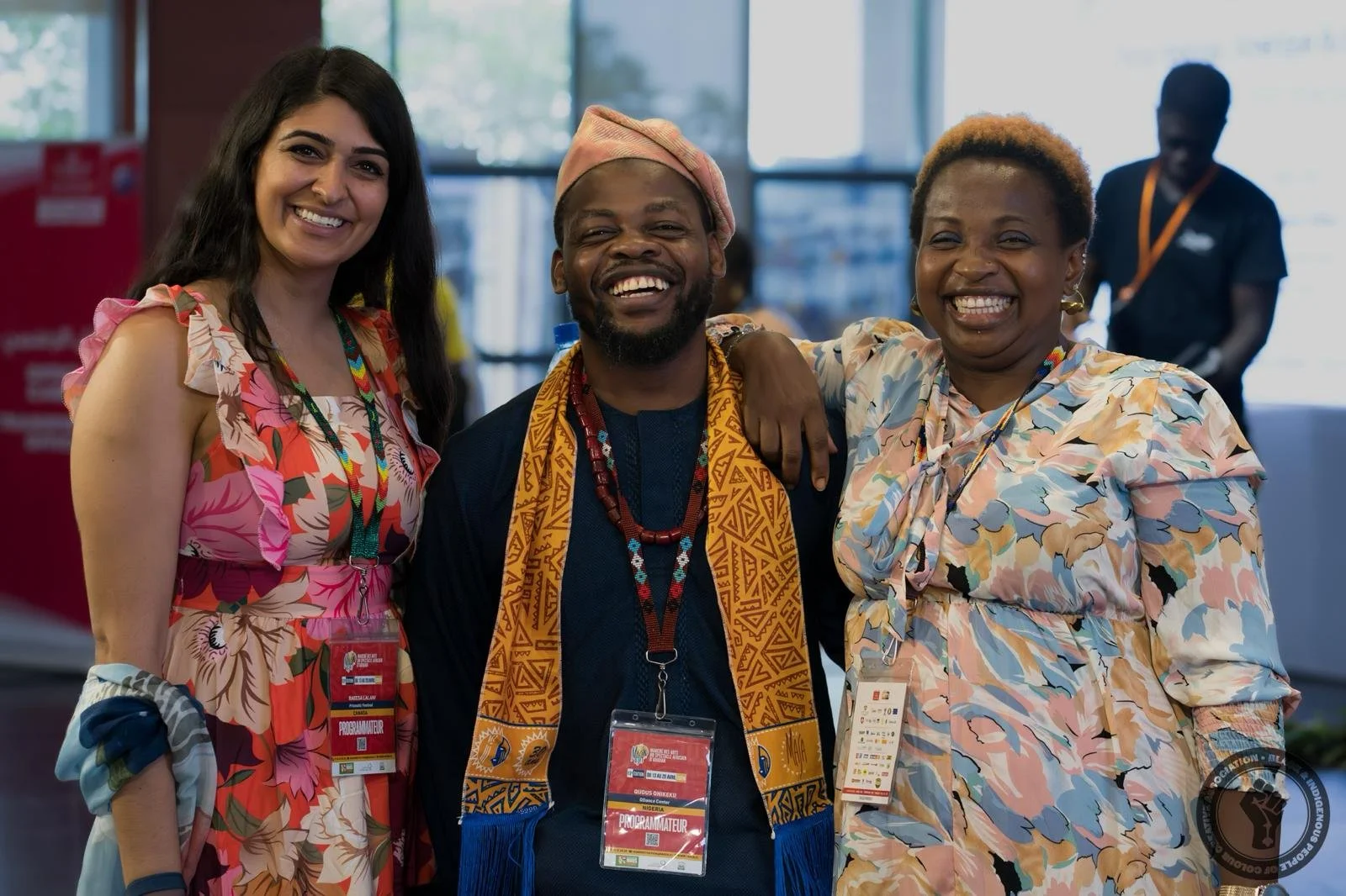 Three people smiling at a conference or event, wearing badges, standing indoors with large windows in the background.