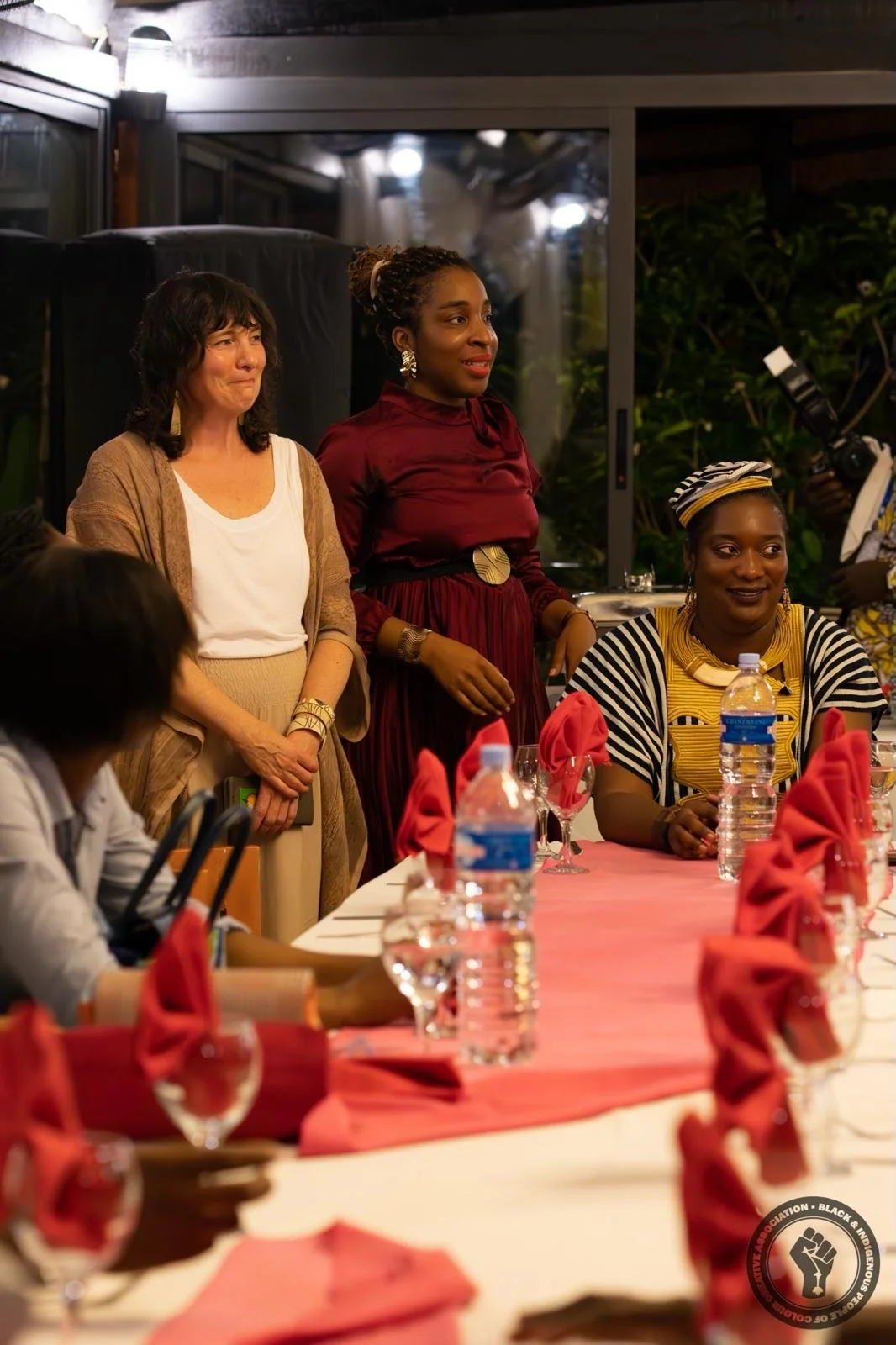 Three women standing and sitting at a table during a formal event or dinner. The woman on the left has dark curly hair and is wearing a beige and white outfit. The woman in the middle has braids, wearing a red dress with a gold belt, and the woman on the right is wearing traditional African attire with a headwrap. The table is set with pink napkins, water bottles, glasses, and utensils.