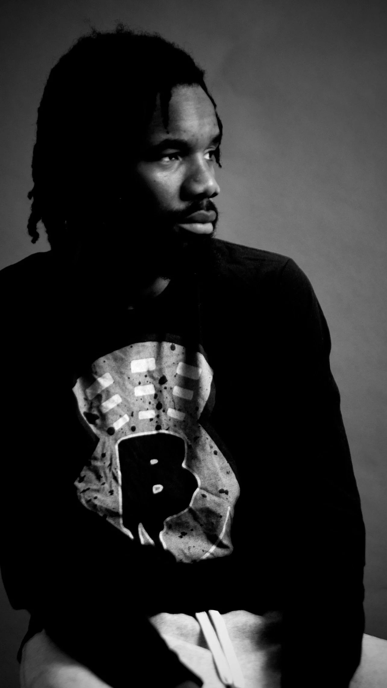 A young man with dreadlocks and a beard, wearing a black sweatshirt with a skull design, sitting with arms crossed, looking to the side in a monochrome portrait.