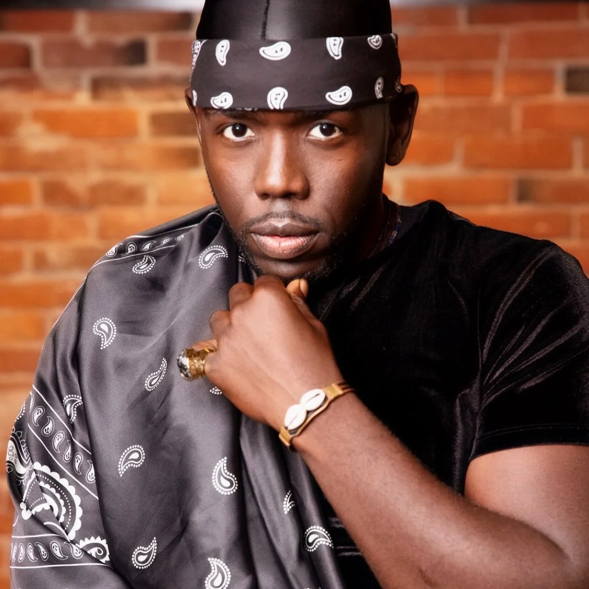 A man with dark skin wearing a black velvet shirt and a black bandana with white paisley patterns. He has a thoughtful expression, resting his chin on his fist, adorned with a gold ring and a bracelet, against a brick wall background.