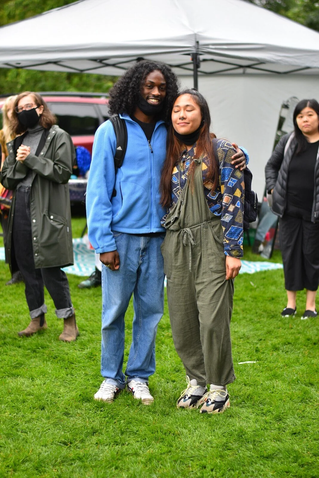 Two people hugging in a park during a gathering, with a tent and other attendees in the background.