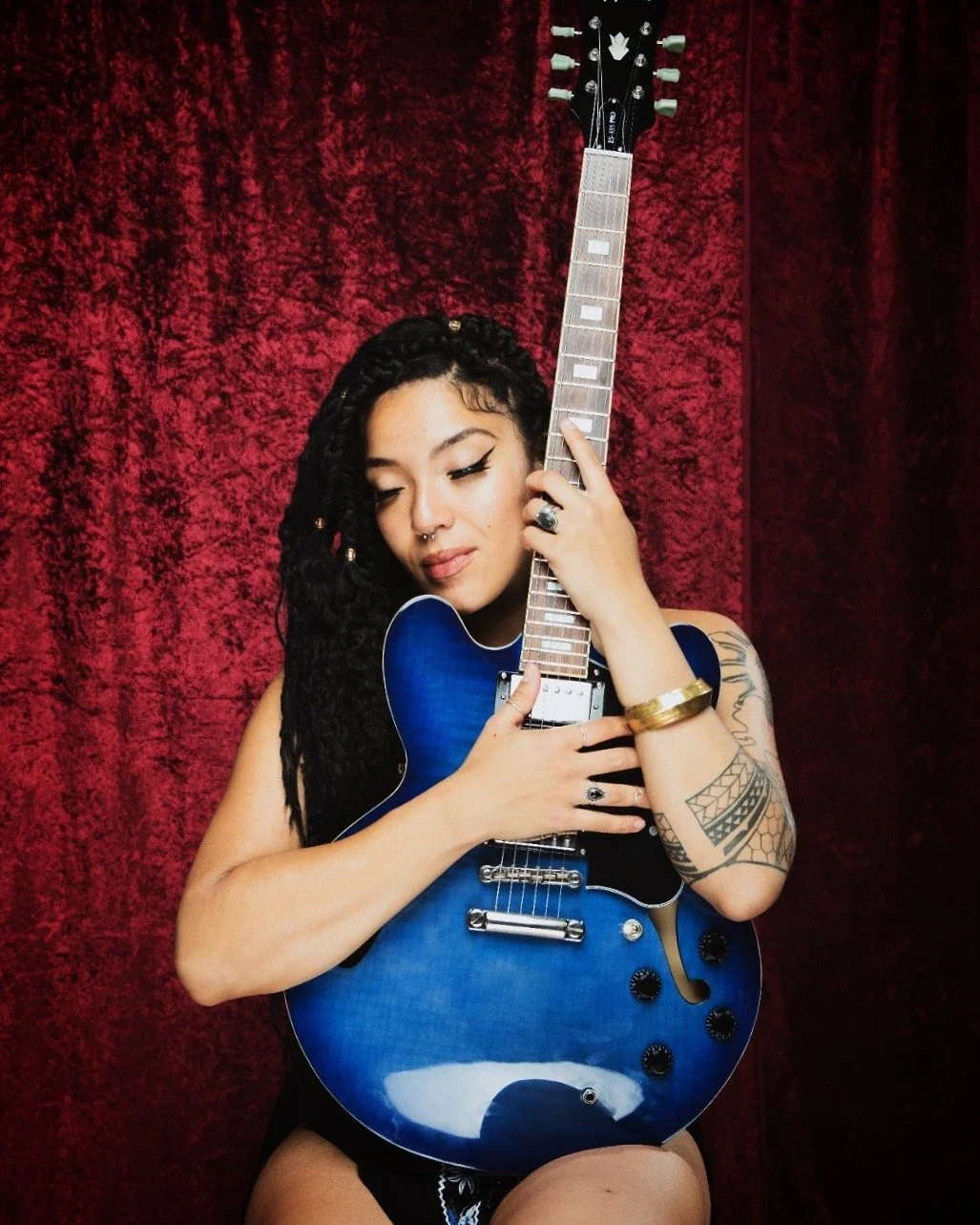 A woman with dreadlocks and tattoos holding a blue electric guitar against a red velvet background.