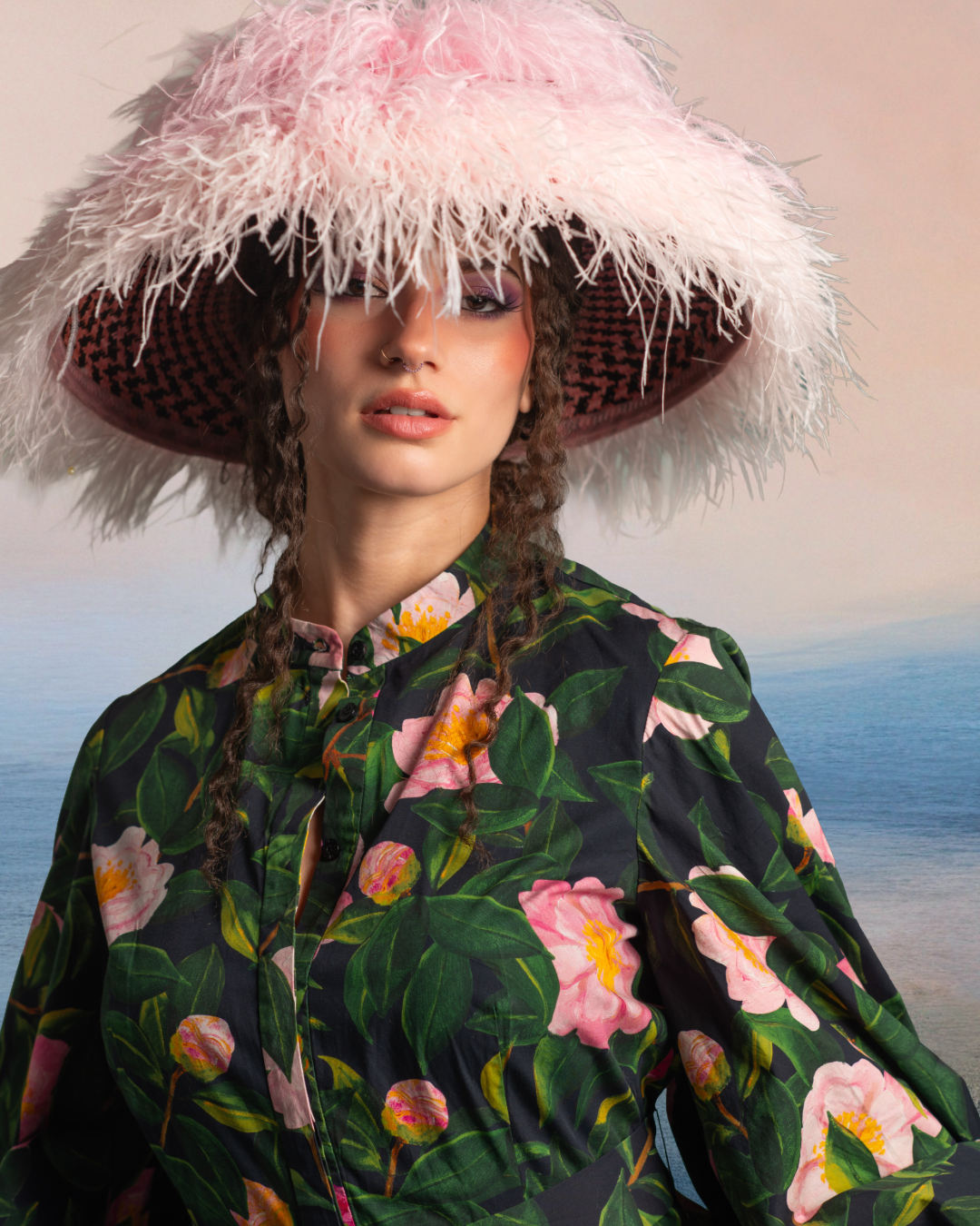 A woman wearing a large pink and white feathered hat with a black and pink checkered underside, a nose ring, and a floral-patterned blouse with green leaves and pink flowers, standing by the ocean during sunset.