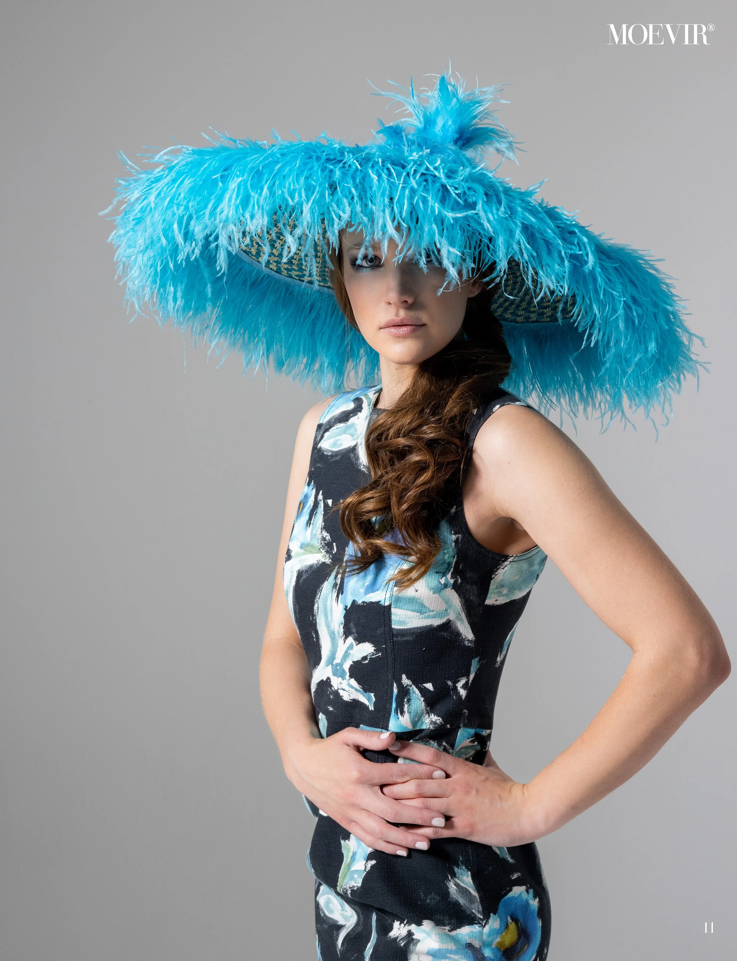 A woman wearing a large, bright blue feathered hat and a sleeveless, patterned dress stands against a gray background, with her left hand on her hip and her hair styled in loose waves.