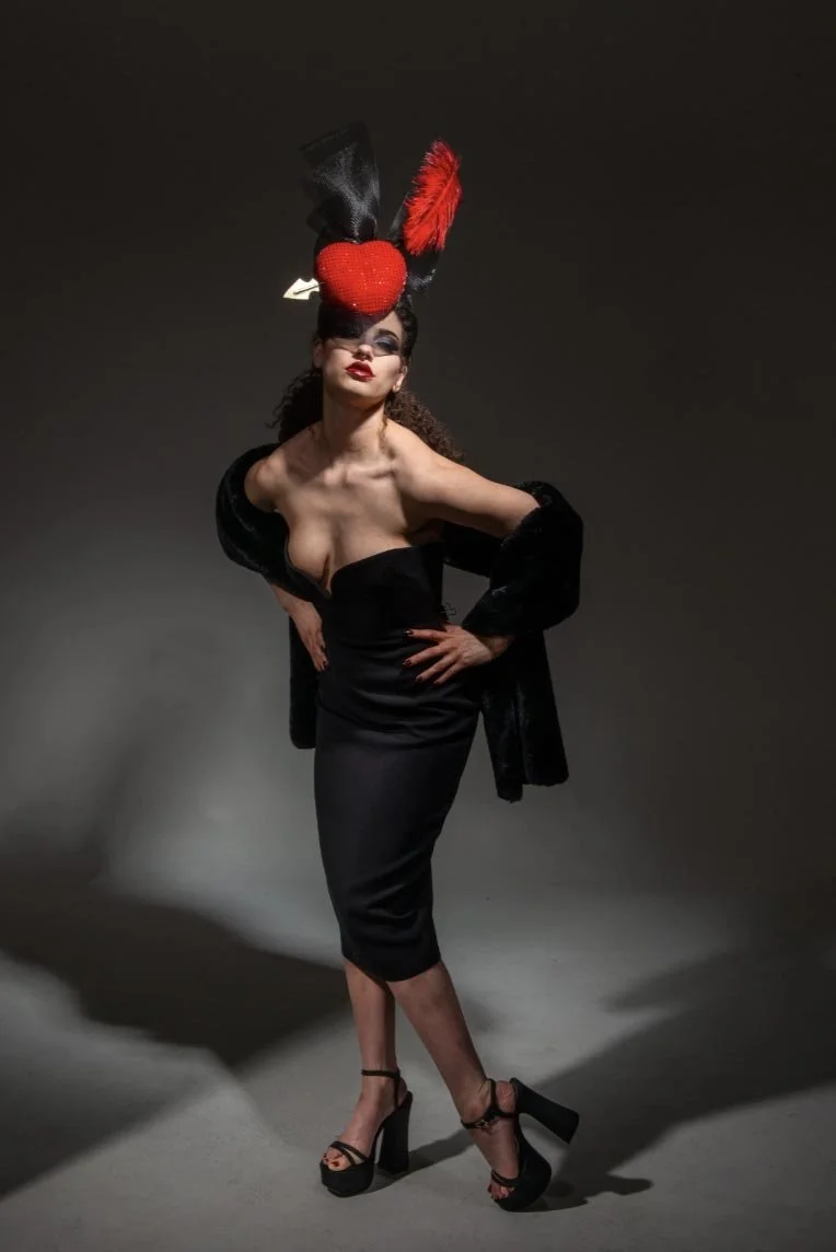 A woman with dark curly hair, wearing a black off-shoulder dress with a slit, black high-heeled sandals, and a dramatic headpiece with a red heart, black tulle, and a red feather, stands with one hand on her hip against a dark background.