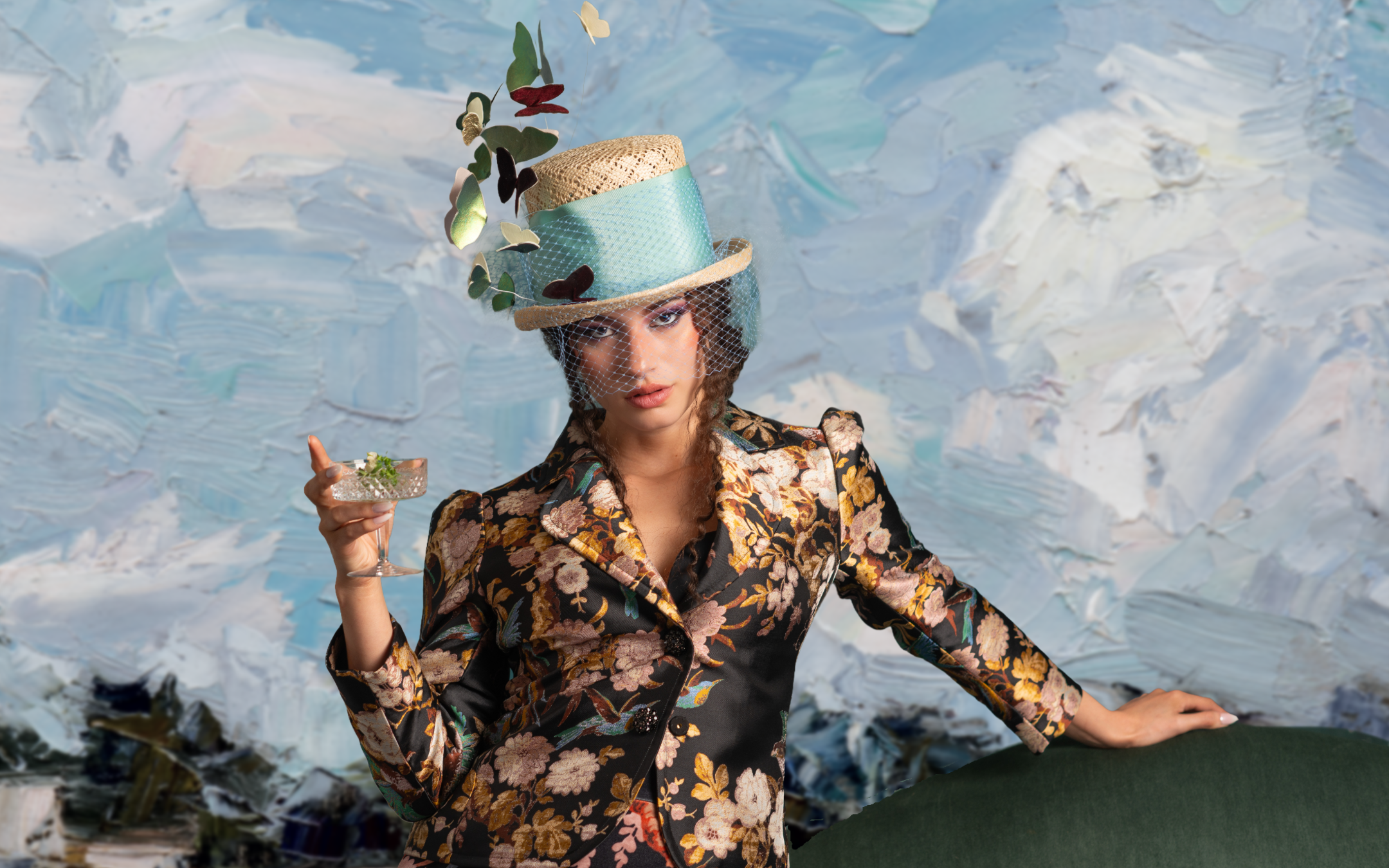 A woman wearing a floral blazer with a mesh hat decorated with butterflies and leaves, holding a cocktail glass, standing in front of an abstract, painted background.