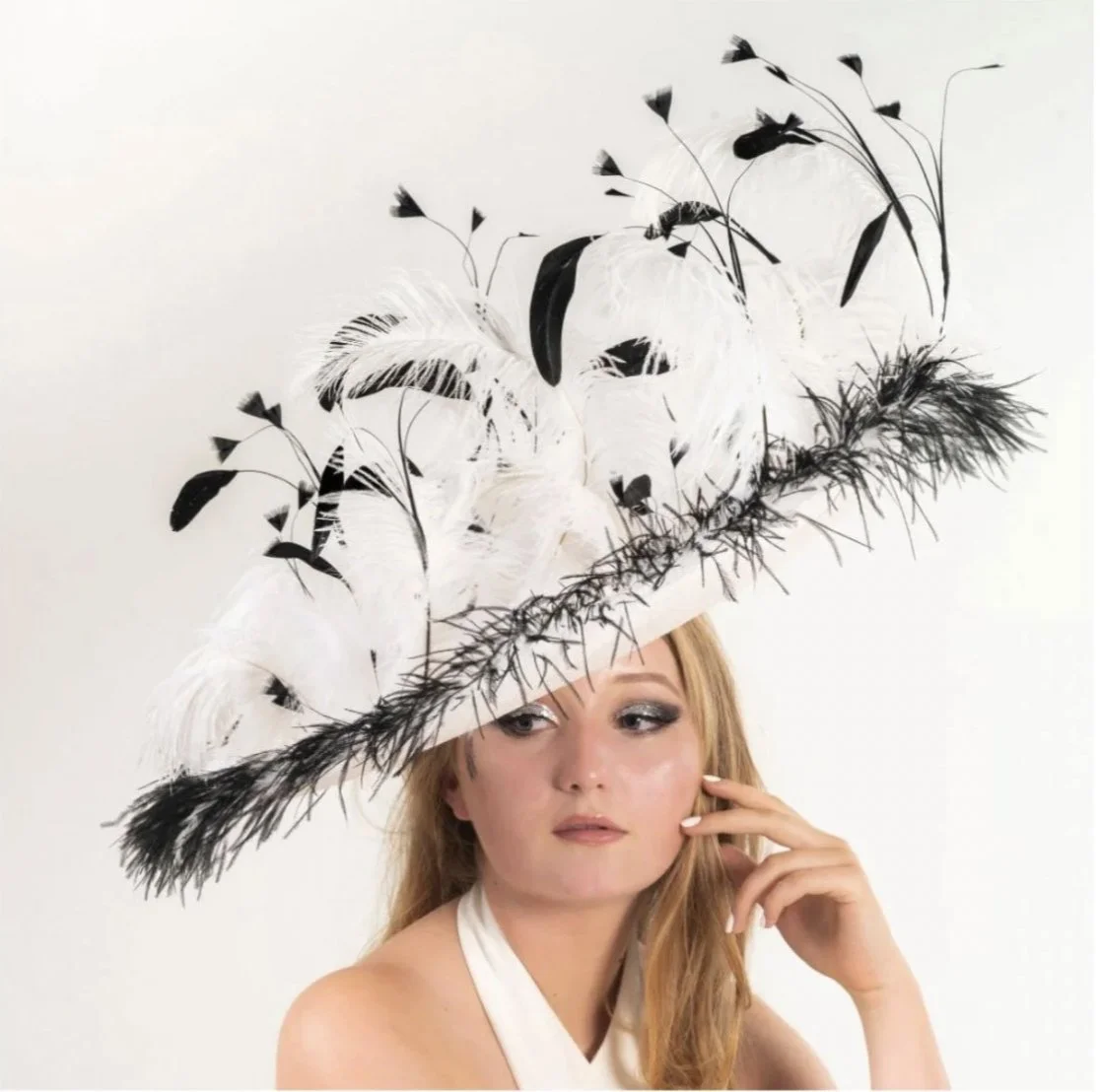A woman wearing an elaborate hat decorated with black and white feathers and floral designs, touching her face with her hand, against a plain white background.