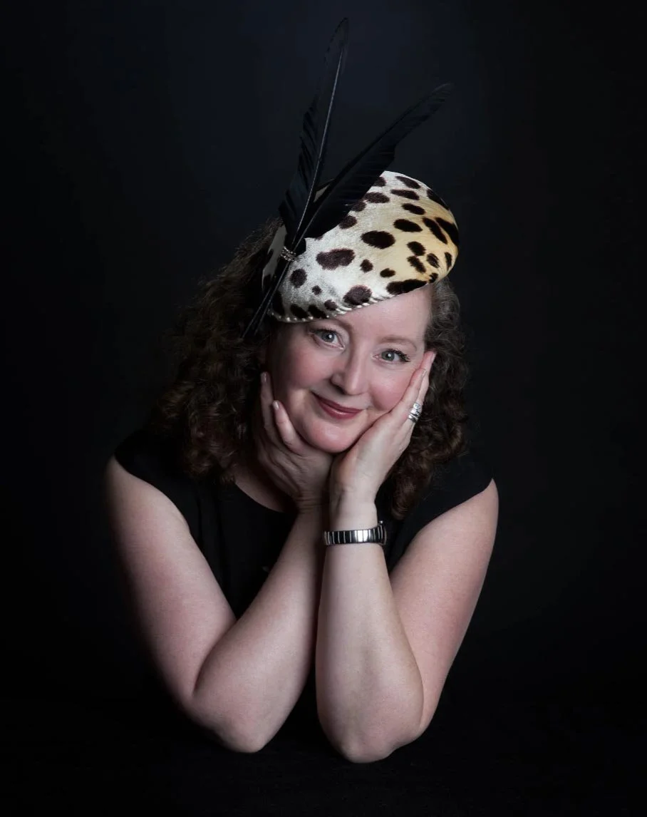 A woman with curly hair smiling and resting her face in her hands, wearing a beige hat with black spots and large black feathers, against a dark background.