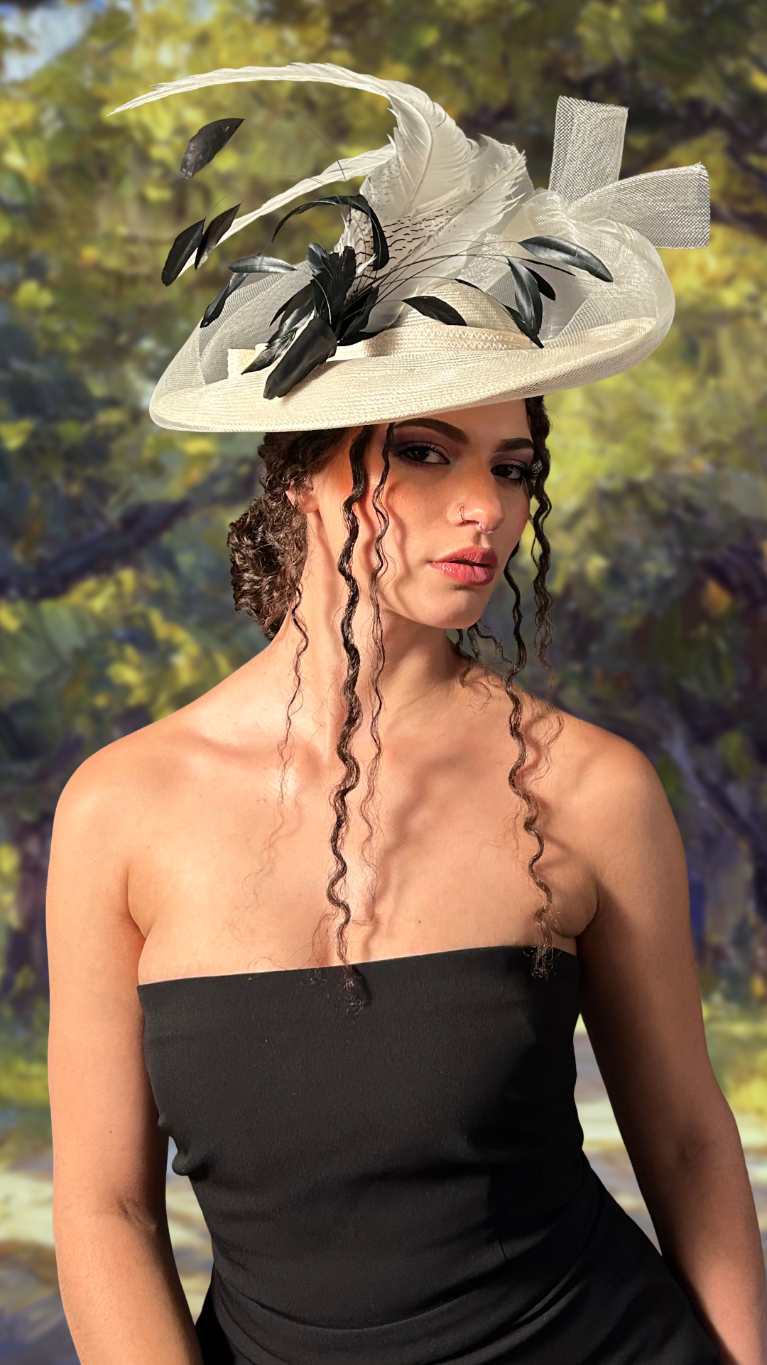 A woman with blonde hair wearing a beige suit with a belt, and a large hat decorated with feathers and animal print fabric, standing outdoors in a garden with trees and a park bench in the background.