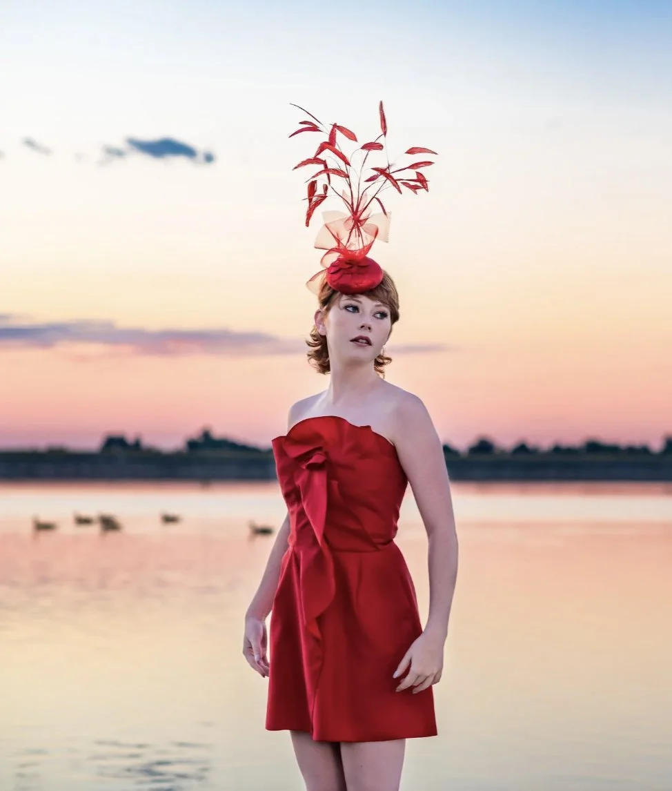 A woman in a strapless red dress and a large red headpiece stands by a body of water during sunset.