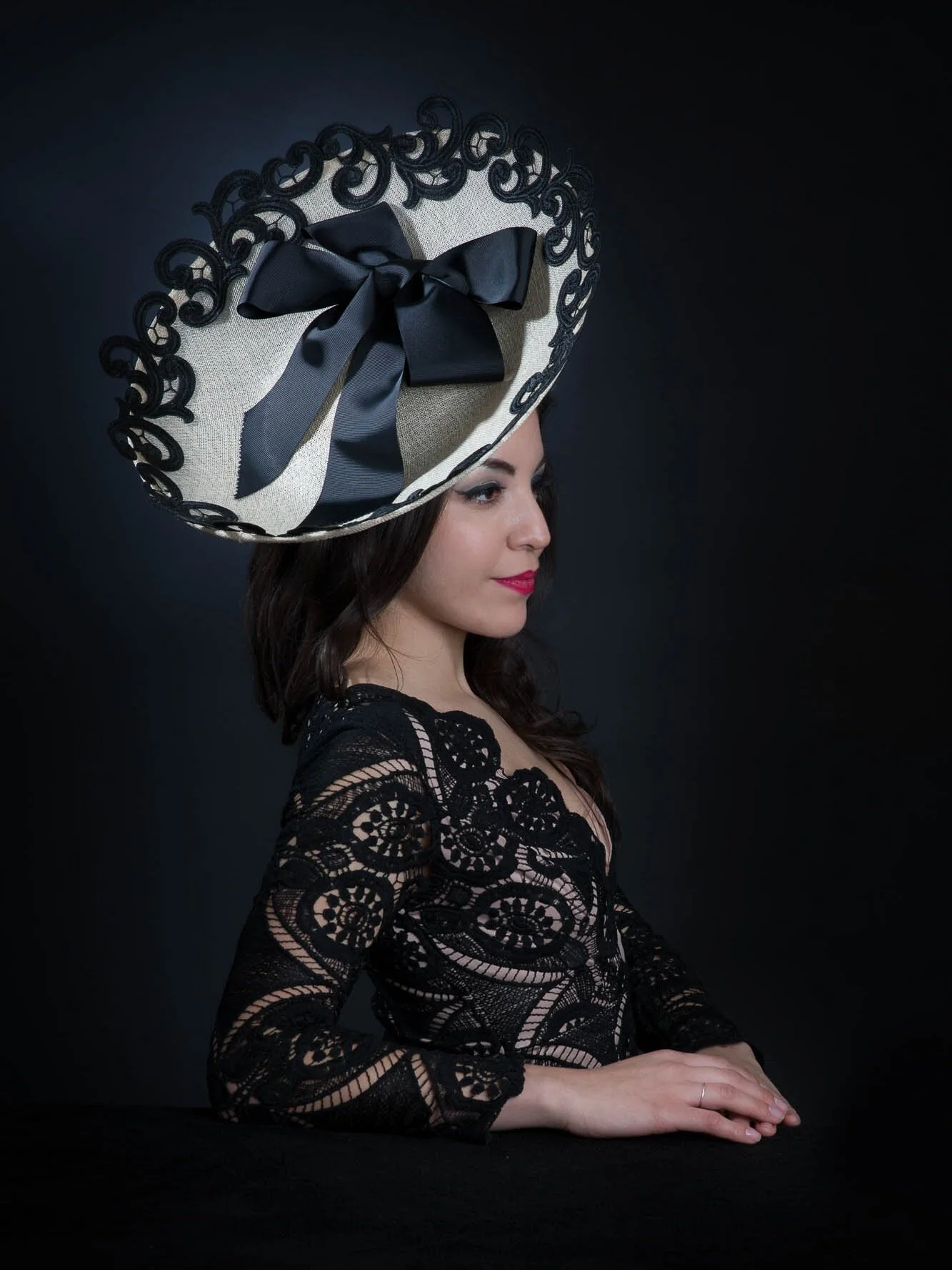 A woman with dark hair wearing a black lace dress and red lipstick, sitting at a table with her hands folded, wearing a large beige hat with black lace and ribbon details, against a dark background.