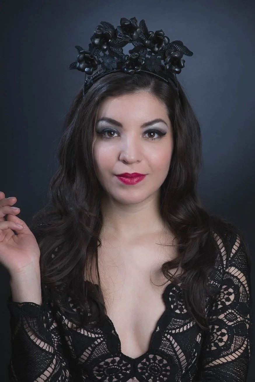 A woman with long dark wavy hair wearing a black lace top and a black floral headband. She has makeup with smoky eyes and red lipstick, and is posing against a dark background.