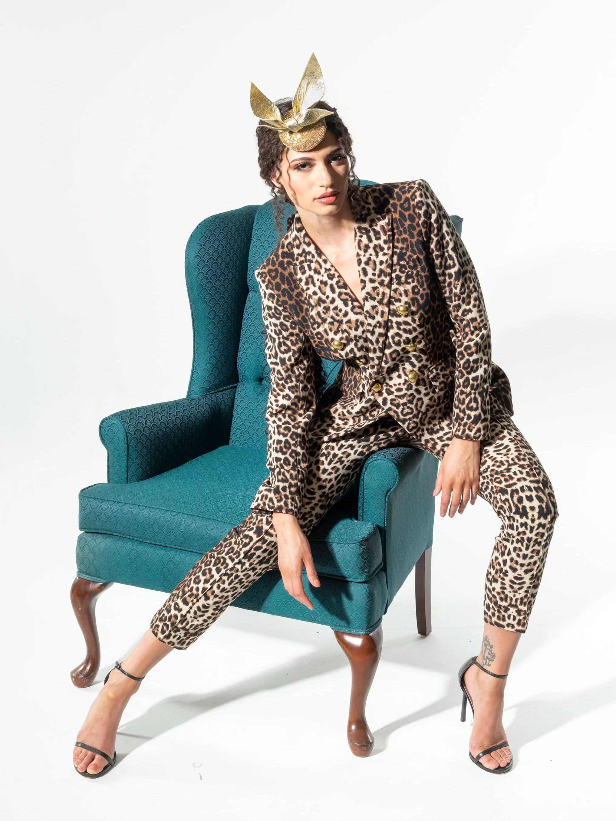 A woman sitting on a teal armchair with leopard print pajamas, wearing black high heels, and a gold headpiece.