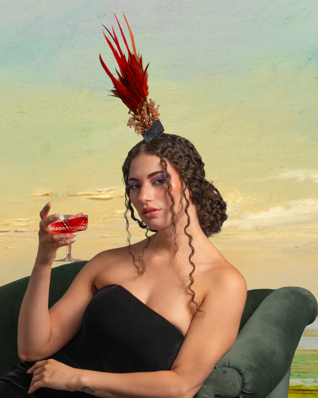 A woman with curly hair sitting on a green sofa, wearing a strapless black top, holding a red cocktail glass, with a decorative feathered headpiece against a pastel-colored background.