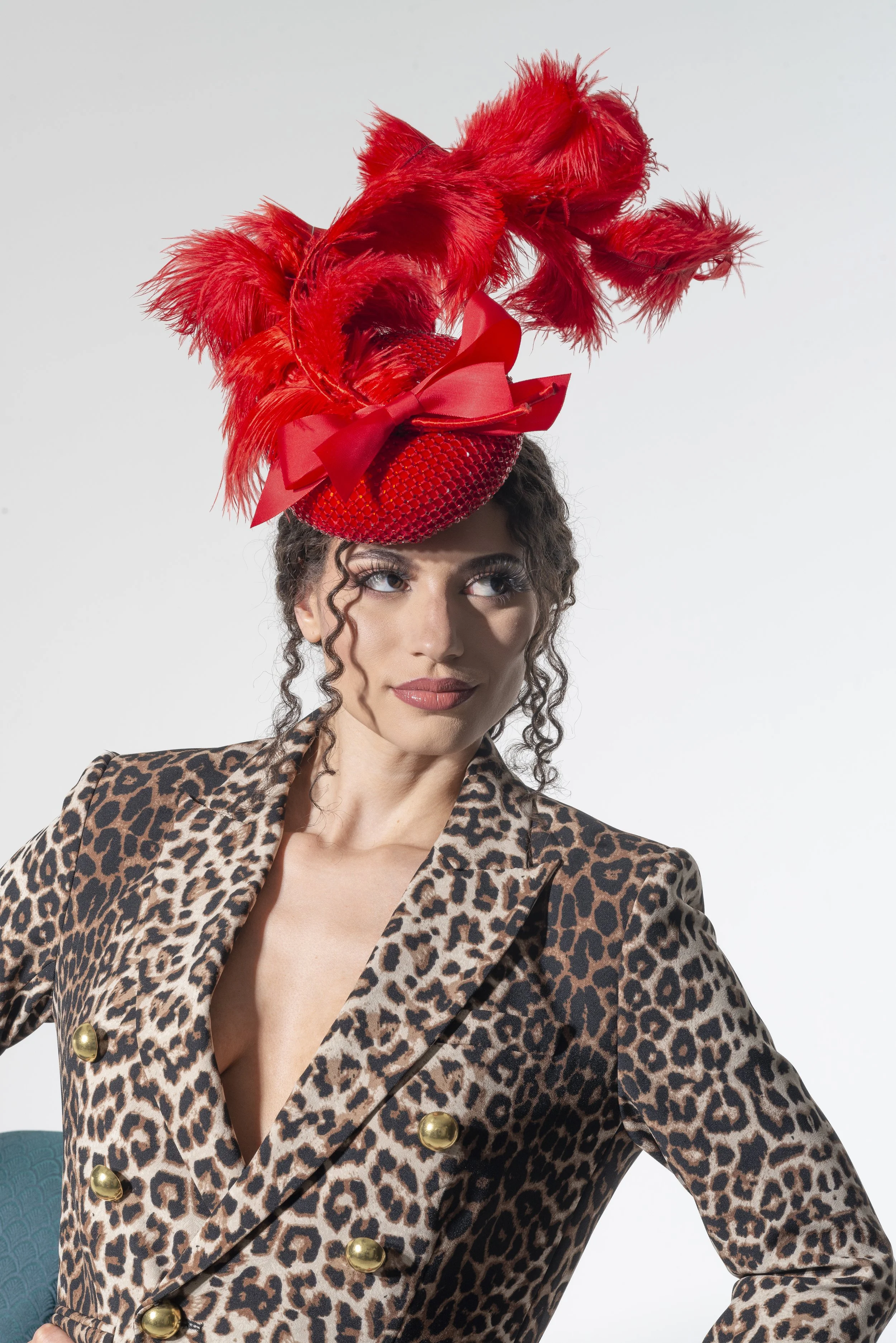 Woman wearing a leopard print blazer with gold buttons and a large red feathered hat with a bow.