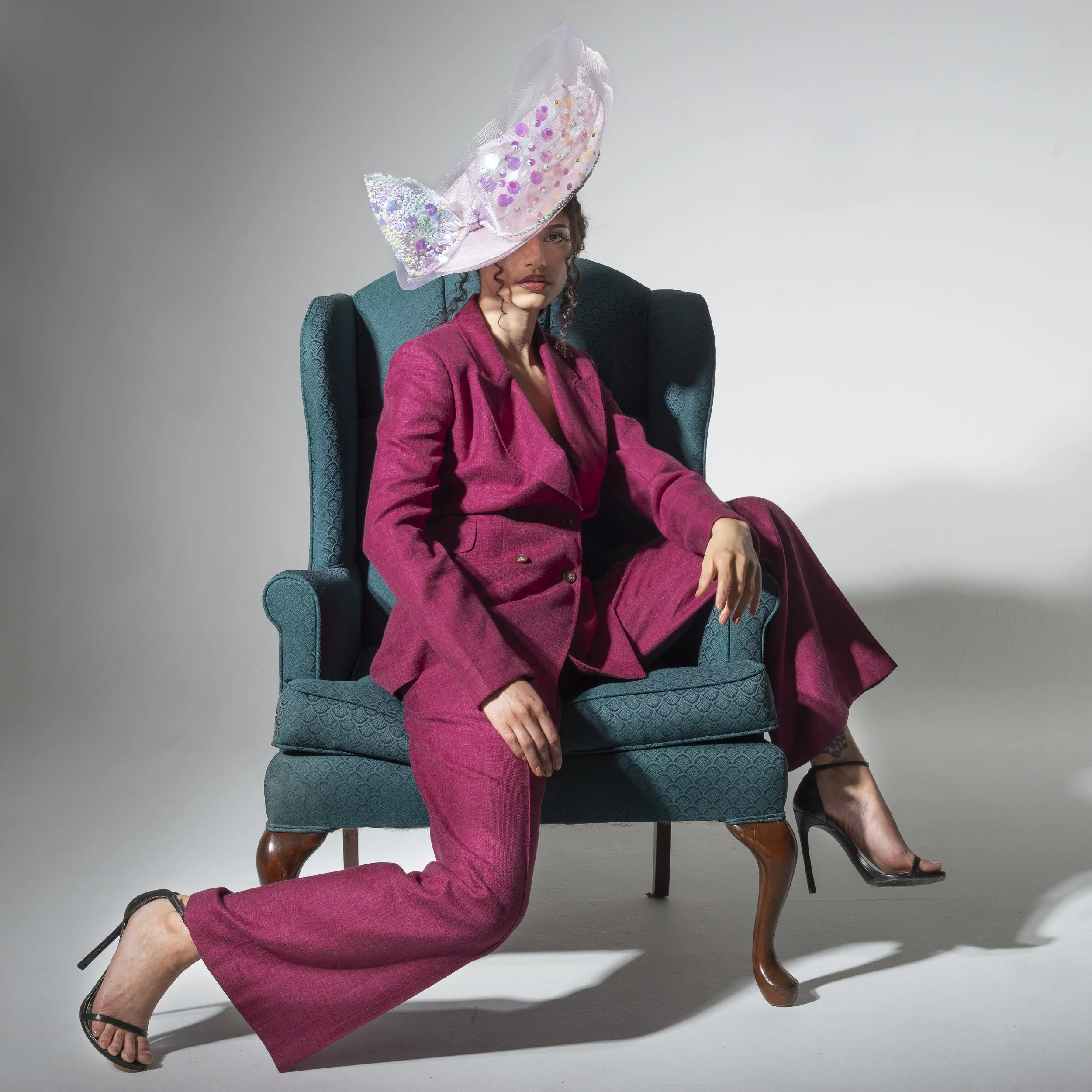 Woman sitting in a teal armchair, wearing a pink suit with a matching pair of trousers and a blazer, black heeled sandals. She has curly hair, partially covered by a large, elaborate pink hat decorated with purple, pink, and iridescent sequins. One of her eyes is partially covered by the hat, and she has a serious expression. The background is plain and light-colored.