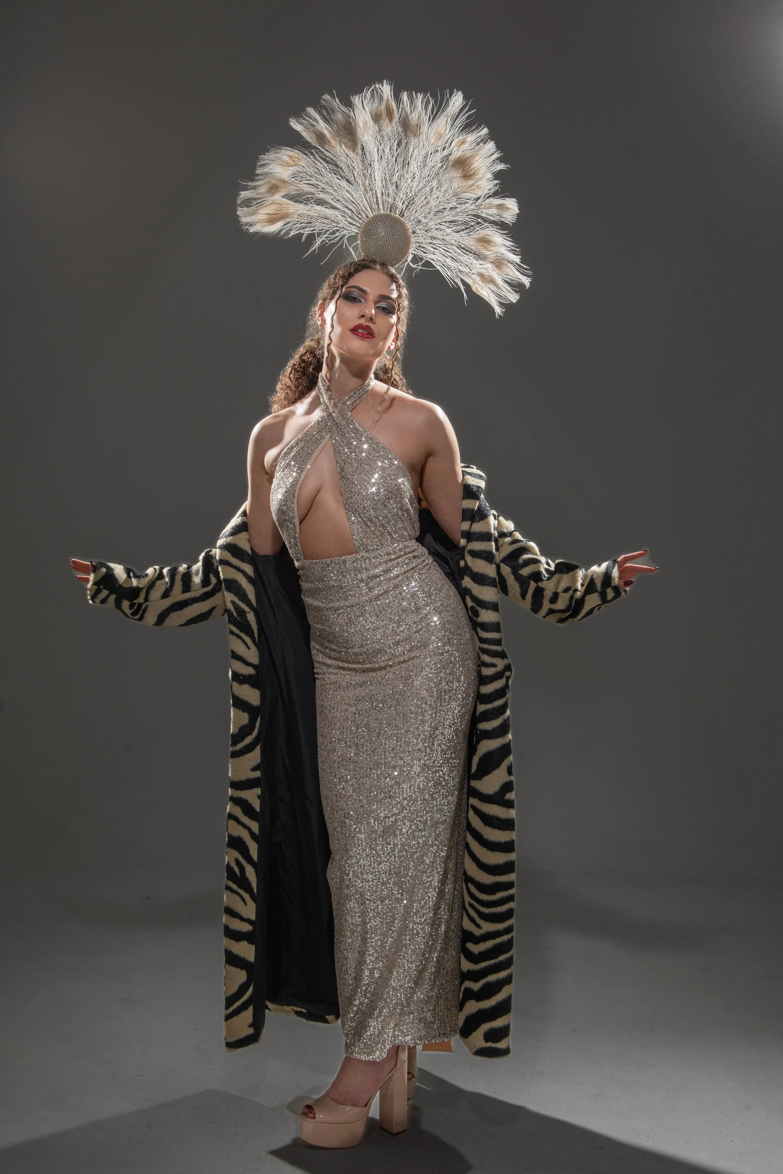 A woman wearing a sparkling, silver, halter neck evening gown with cut-out details, paired with nude high heels, and a zebra-print fur coat, stands against a plain gray background. She has curly hair, bold makeup with red lipstick, and is accessorized with earrings and a choker.
