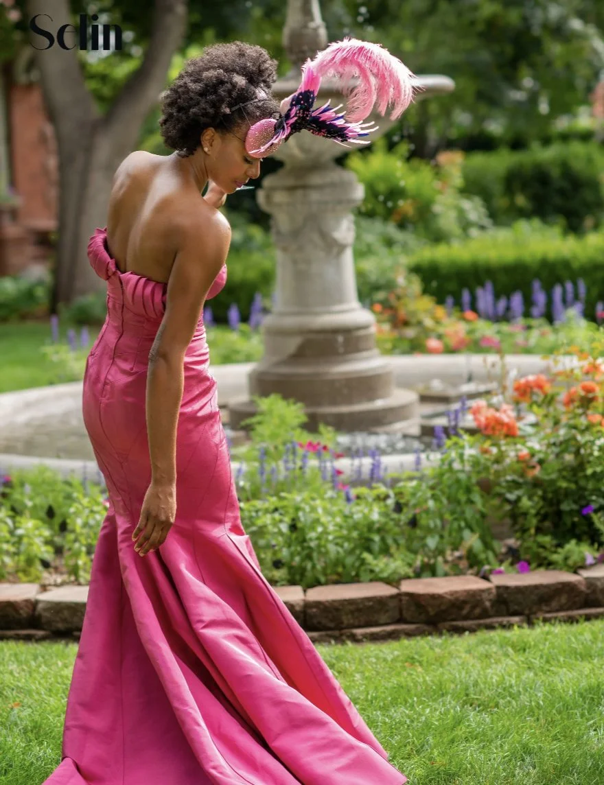 Woman in a strapless pink gown with a large pink feathered headpiece standing in a garden.
