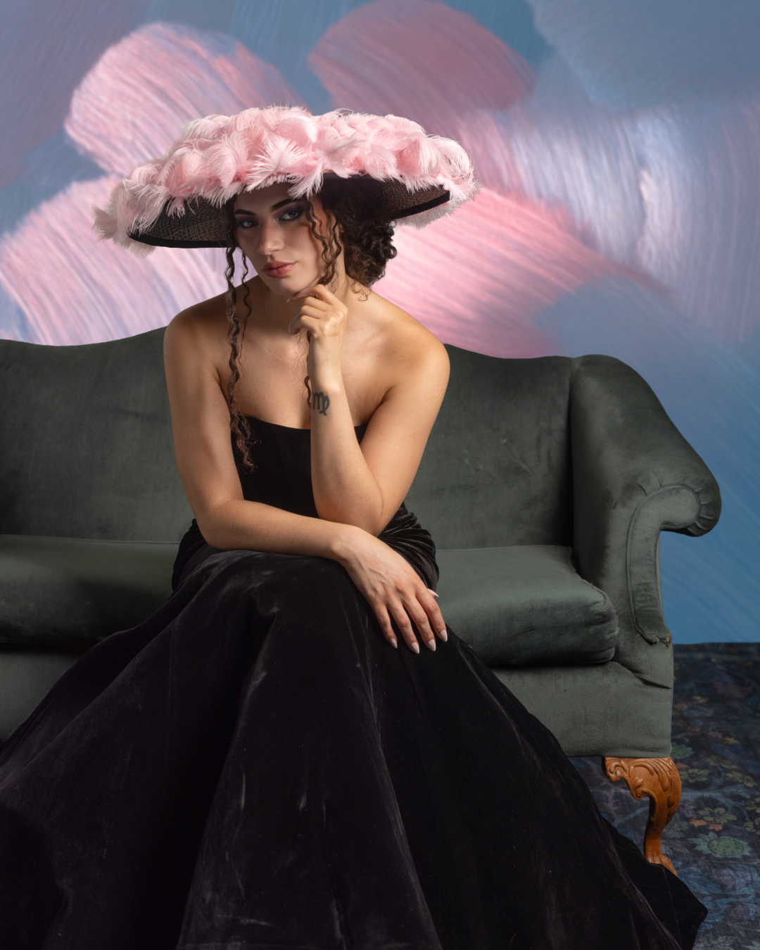 A woman wearing a large pink feathered hat, a strapless black dress, sitting on a vintage green sofa in front of a colorful abstract painted wall.