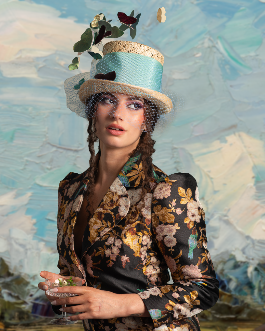 A woman wearing a floral blazer and an elaborate hat with greenery, lace, and fabric decorations, holding a glass dish with small plants, against an abstract, textured background.
