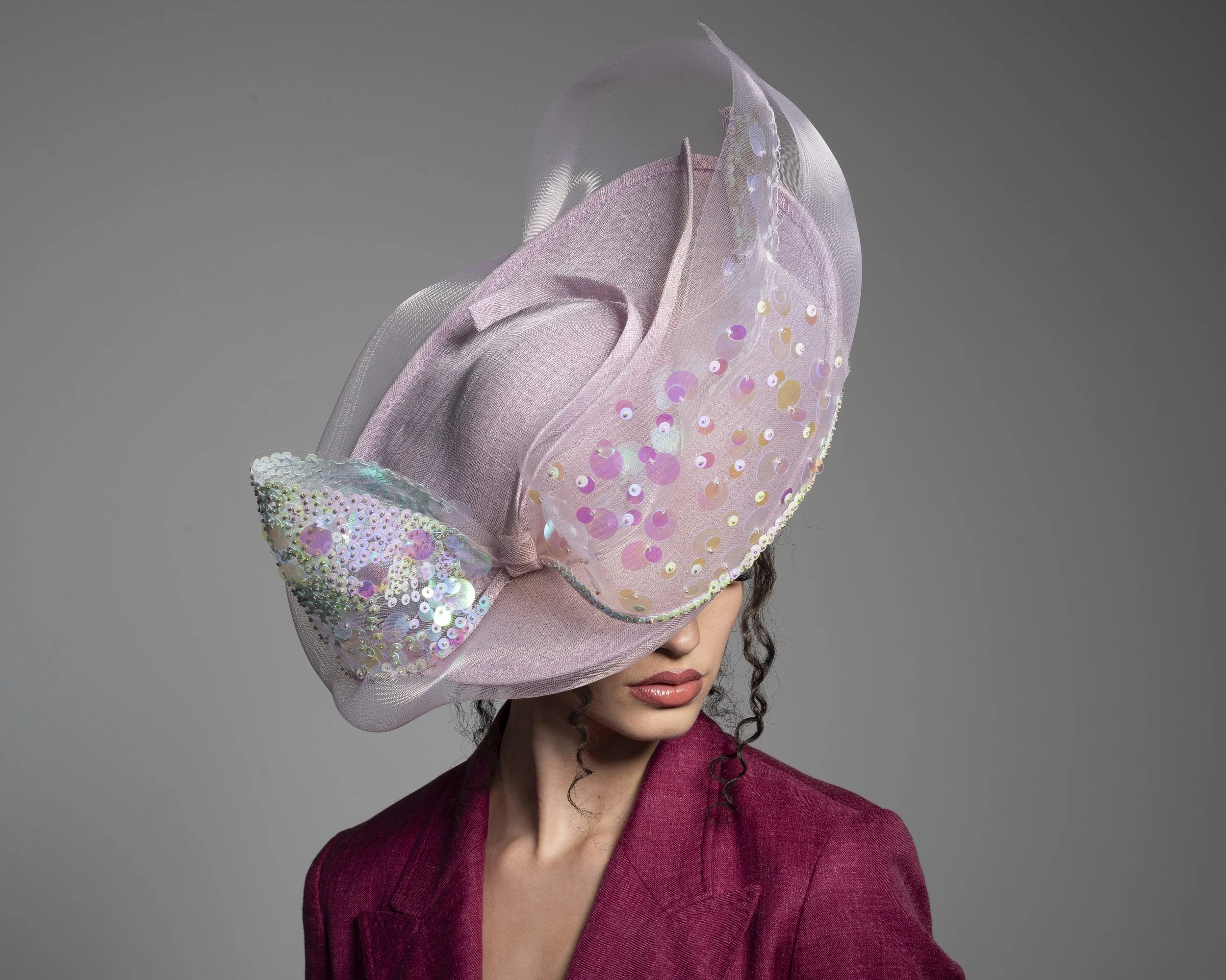 A woman with dark, curly hair wears a pink fascinator with iridescent sequins and a netted veil, and a matching pink blazer, set against a plain gray background.