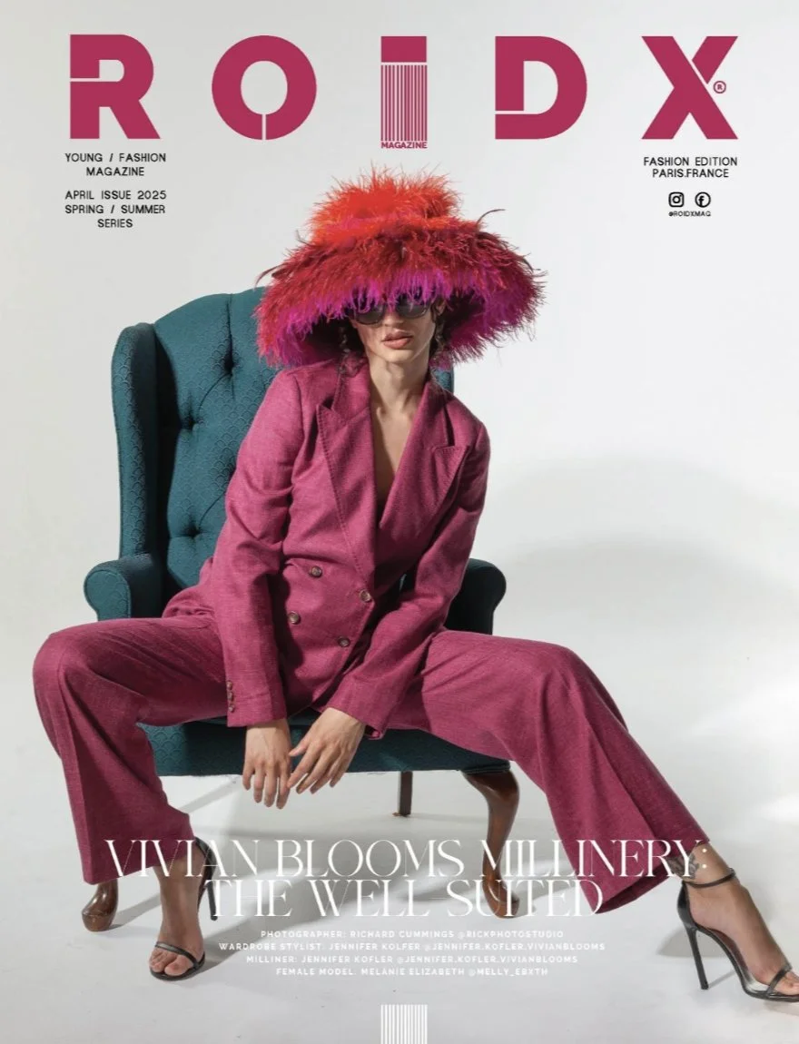 Fashion magazine cover featuring a woman in a pink suit with a large colorful feathered hat sitting on a teal armchair.