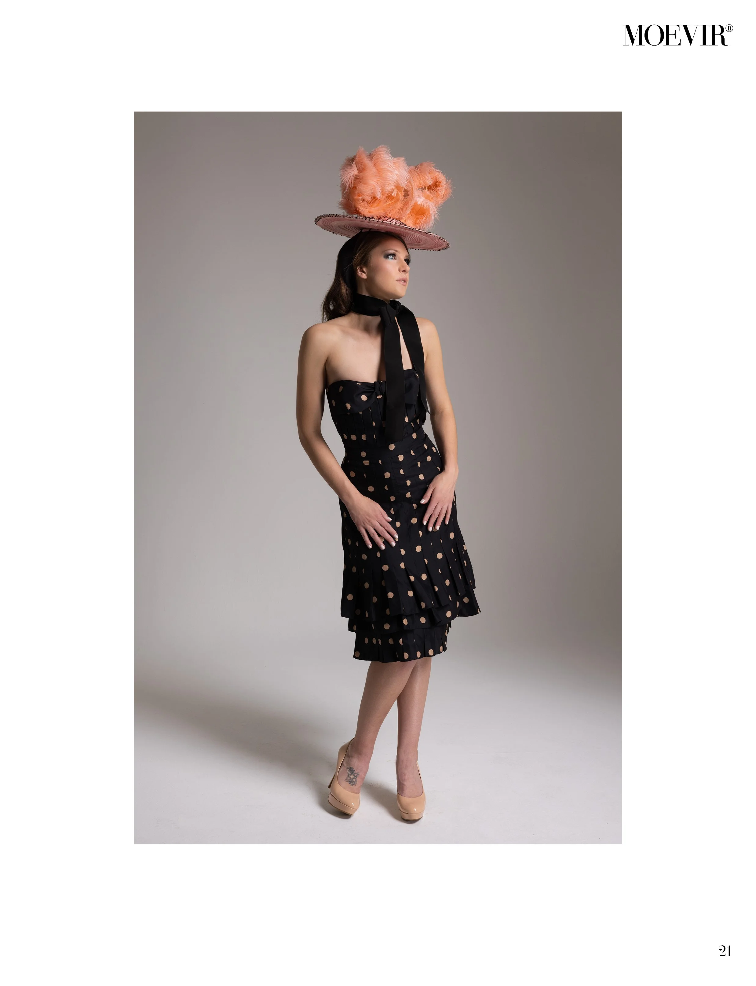Fashion model wearing a black strapless polka dot dress with ruffled hem, nude heels, black choker, and large pink hat with feathers, standing against plain gray background.