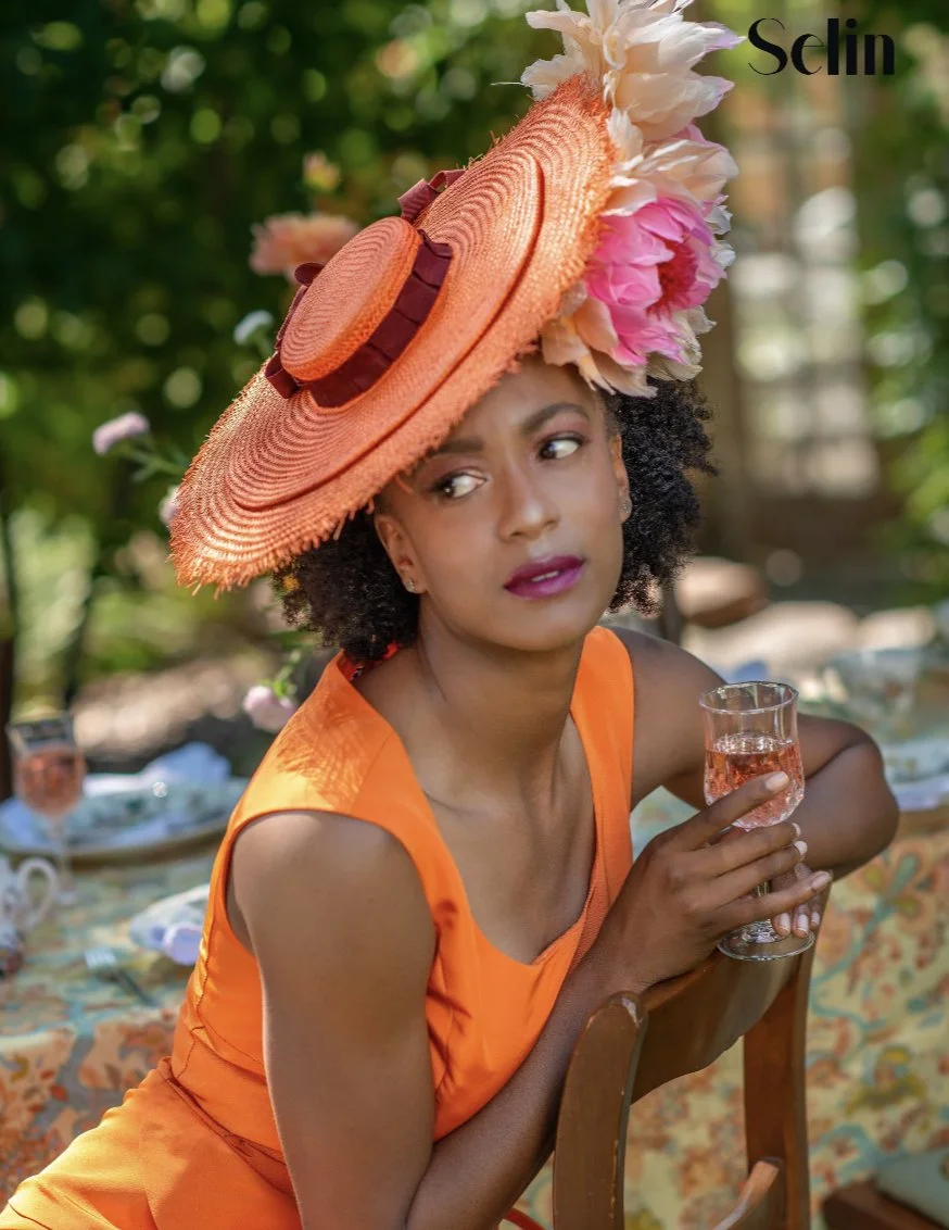 A woman in an orange dress and a large, decorated orange hat with pink and cream flowers, sitting outdoors at a table with a glass of pink drink, surrounded by a garden setting.