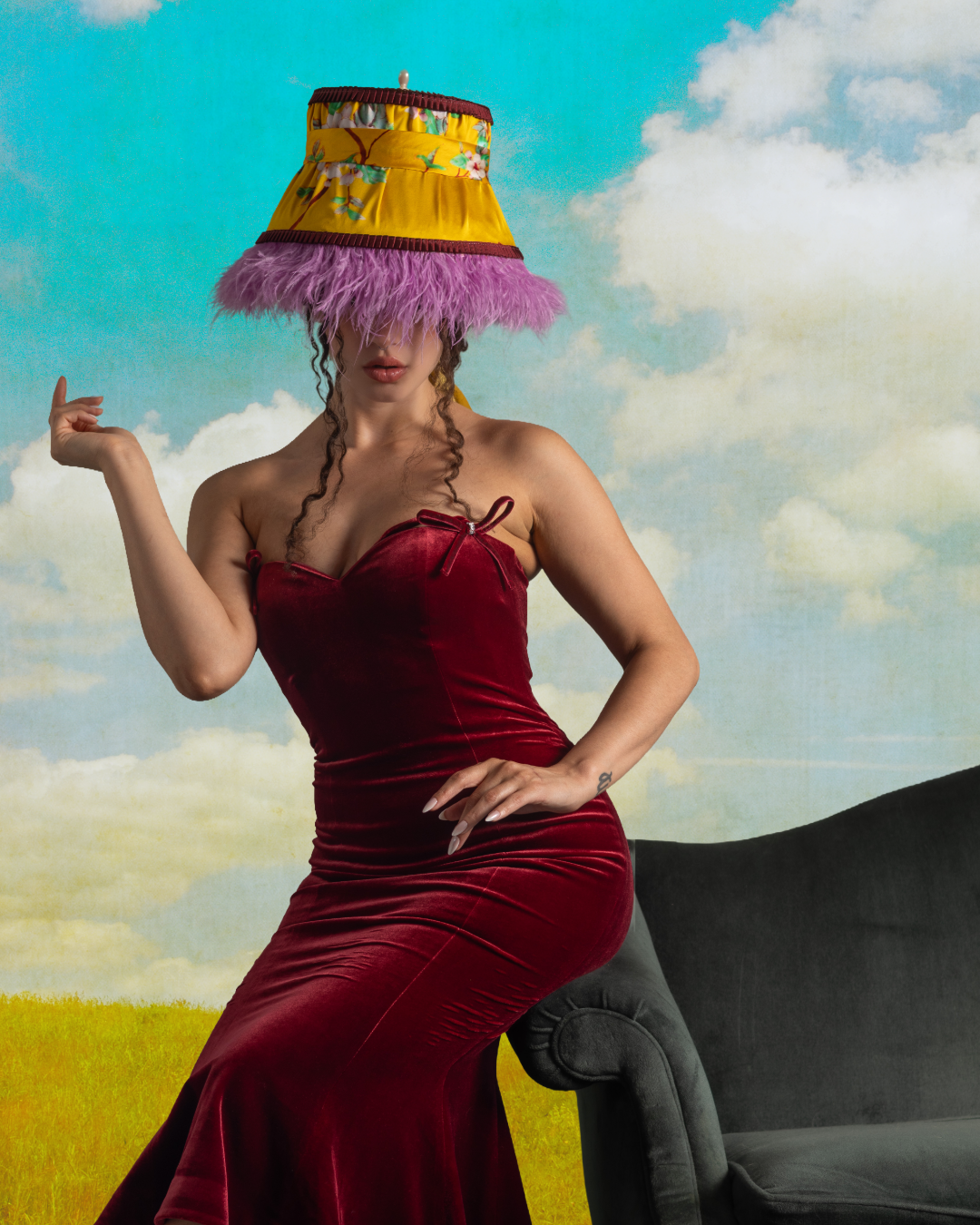 A woman wearing a strapless red velvet dress and a large, colorful hat with pink feathers, standing beside a gray sofa against a sky and grass background.