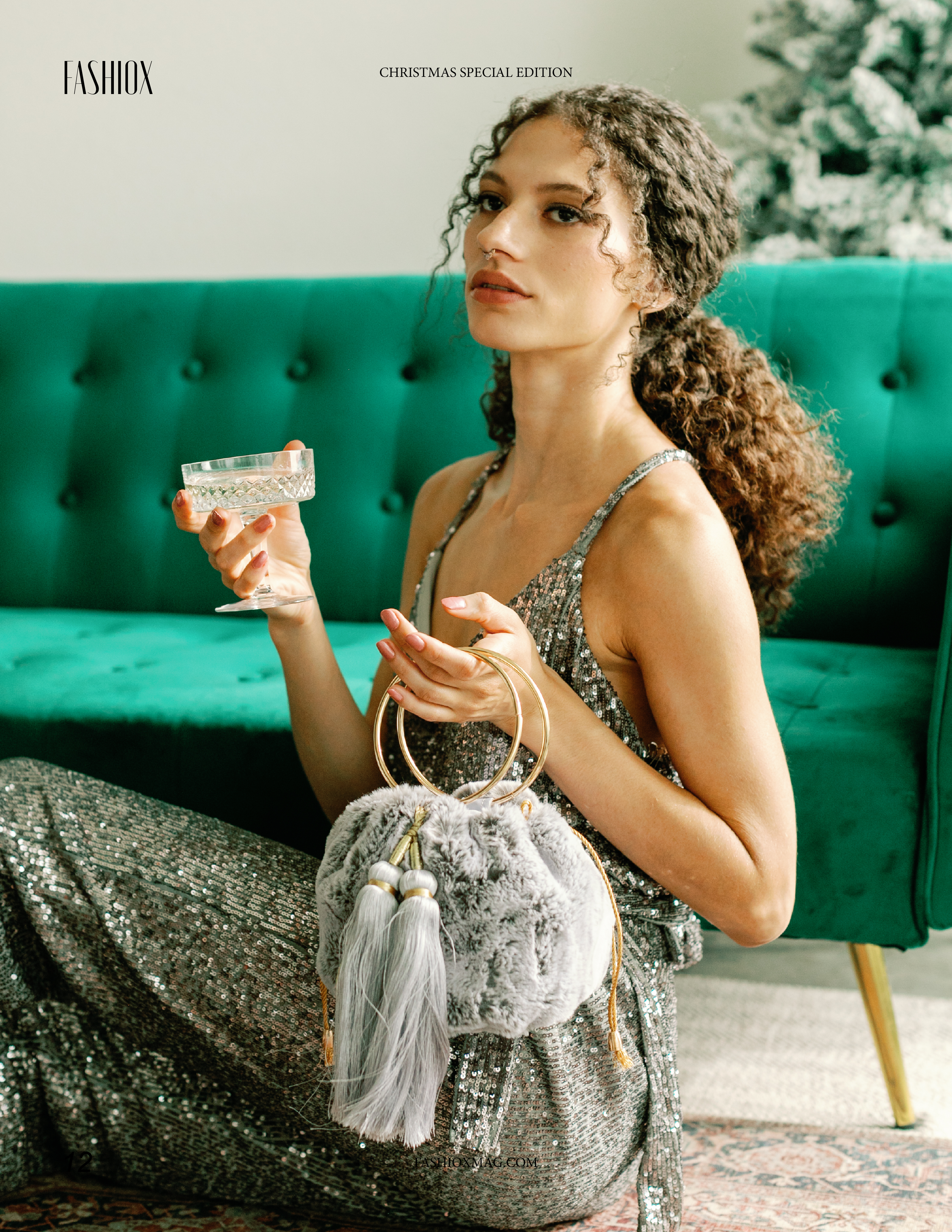 A woman with curly hair sitting indoors on a green tufted sofa, holding a crystal glass with a drink, wearing a sequined dress, and holding a furry bag with a tassel and large gold rings.