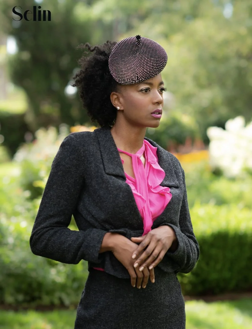 A woman with curly hair in a large beaded hat, wearing a pink blouse, dark gray jacket, and matching skirt, standing outdoors in a garden.