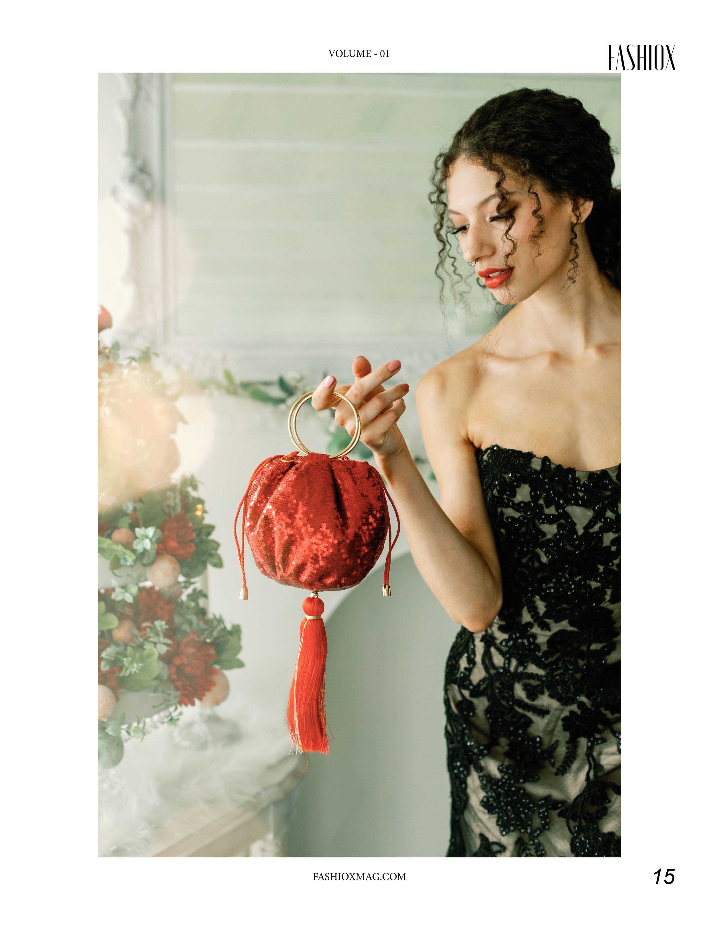 A woman in a black lace dress looking down while holding a red sequined handbag with gold handles and tassels. There is a Christmas tree decorated with white ornaments and flowers in the background.