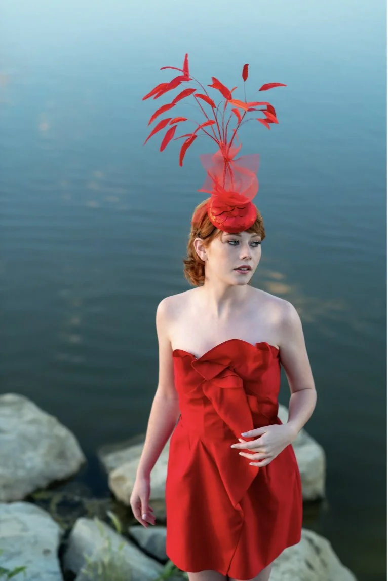 A young woman with short red hair stands by a body of water, wearing a strapless red dress with ruffle details and a large red bow. She has an elaborate red headpiece featuring feathers and tulle.