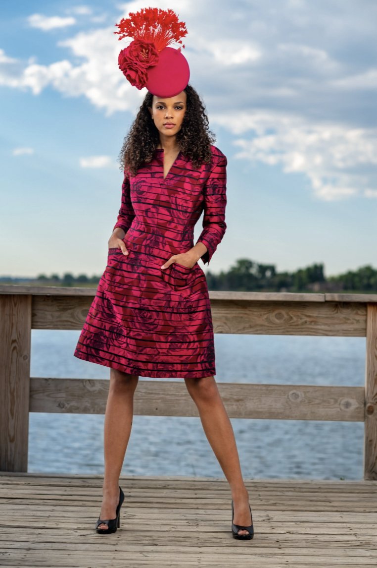 A woman standing on a wooden dock by the water, wearing a red dress with black patterns, and an elaborate red hat with feathers and flowers, against a partly cloudy sky.