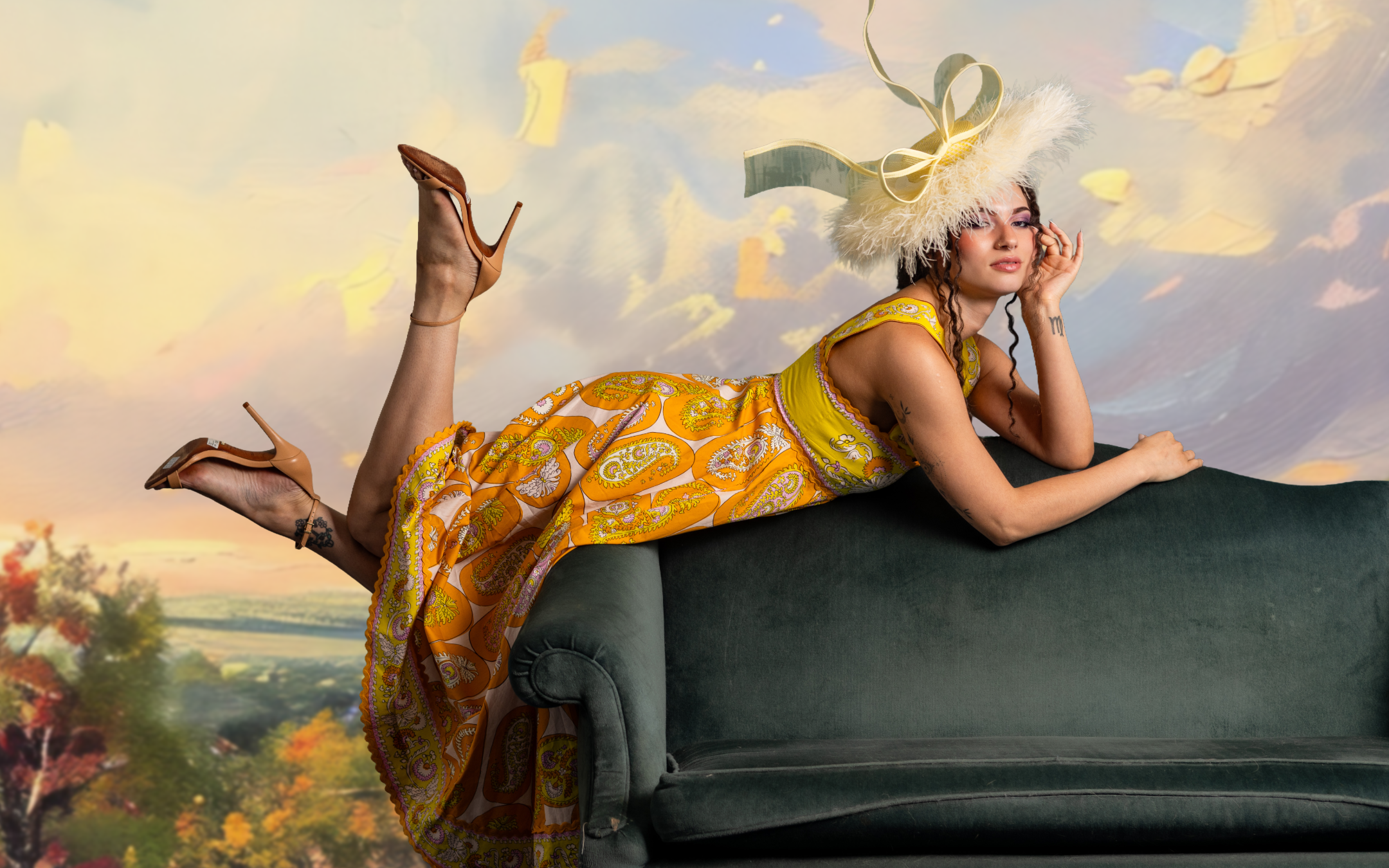 A woman wearing a yellow Indian-style dress and a large feathered hat with ribbons is lying on her stomach on a dark green velvet sofa, with her legs bent at the knees and her feet crossed in the air. The background features a pastoral landscape with trees and a cloudy sky.