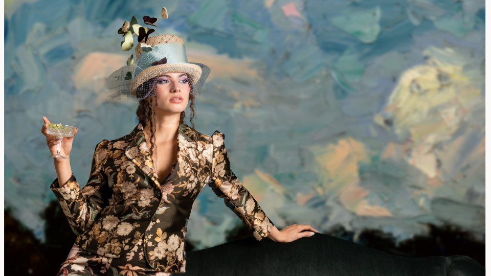 A woman dressed in a floral patterned suit and a decorative hat with flowers and netting, holding a glass of drink, standing in front of an abstract painted background.