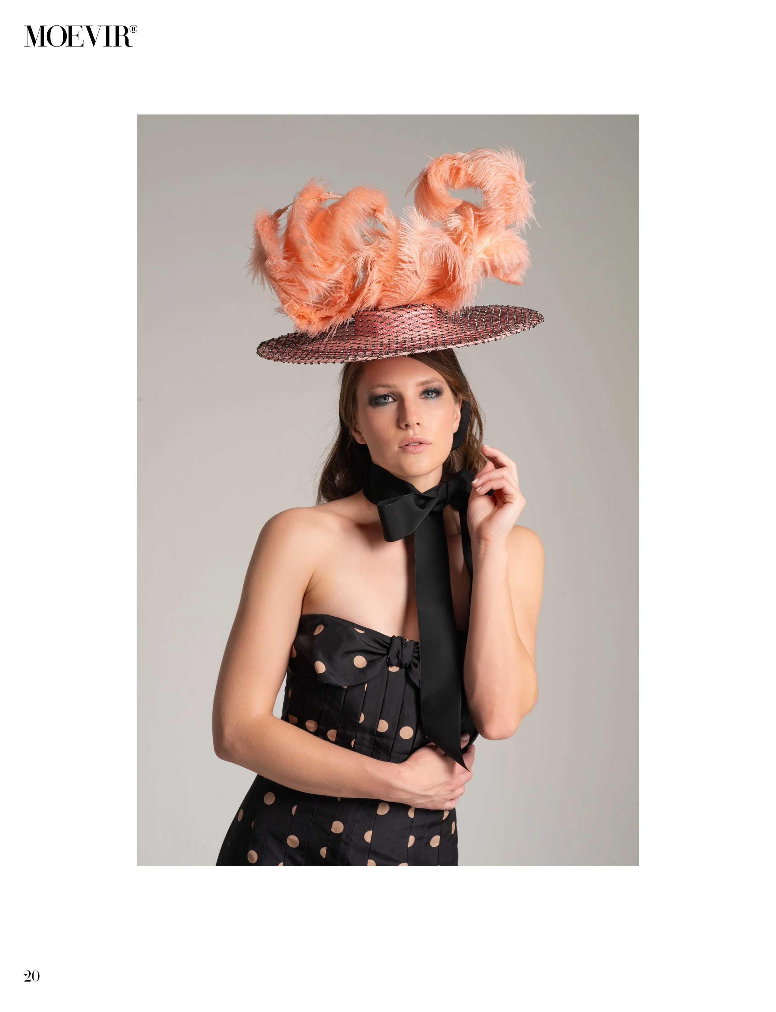 A woman wearing a pink book hat decorated with feathers and a polka-dotted black dress with a bow, standing with her hand on her face.