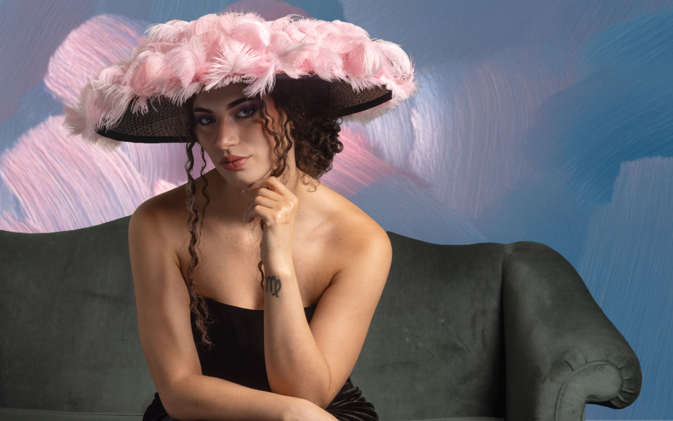 Young woman with curly hair wearing a large pink feathered hat, sitting on a gray couch with a blue and pink abstract background.