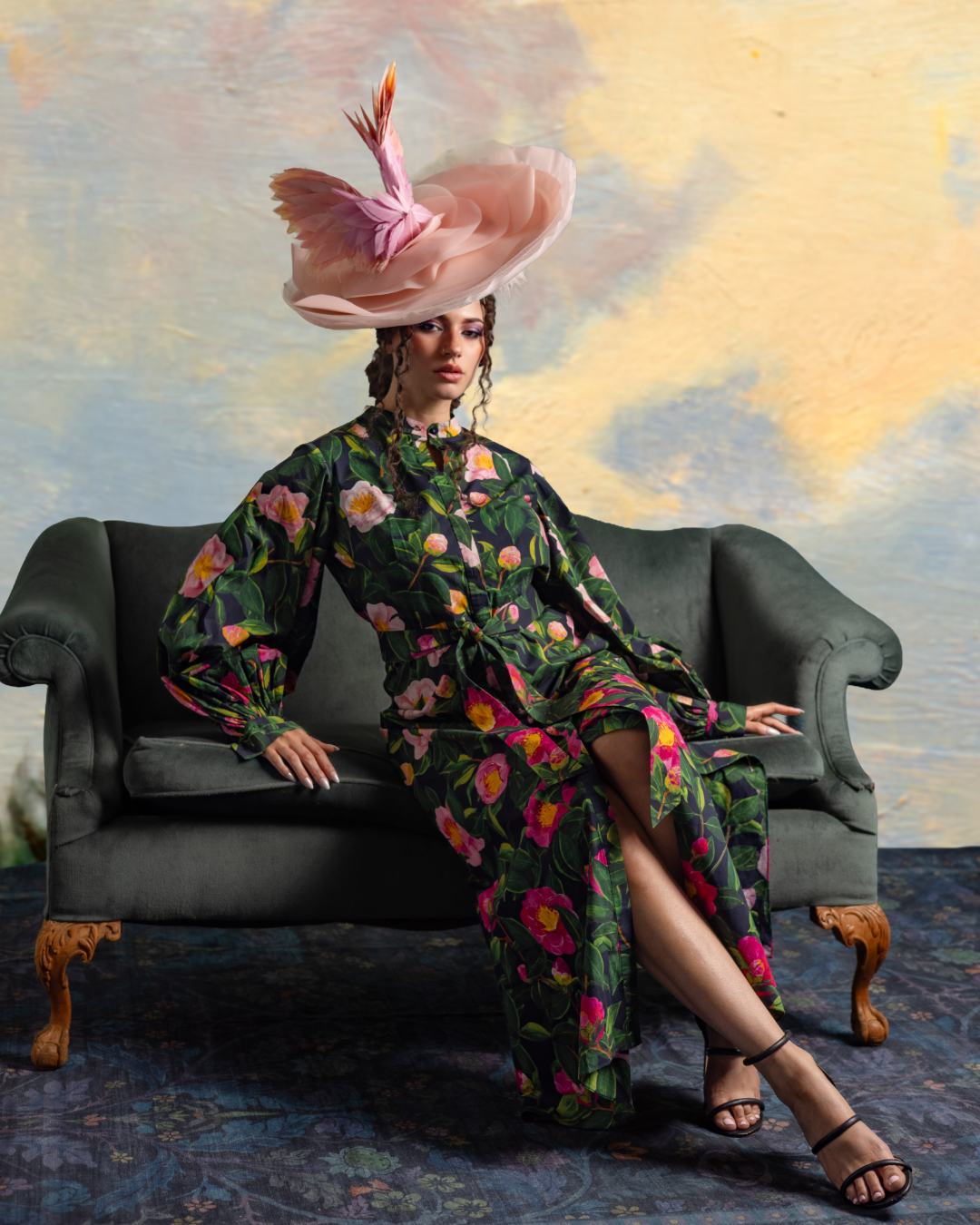 A woman seated on a vintage sofa, wearing a floral patterned dress and a large pink hat with feathers, in front of a painted sky background.