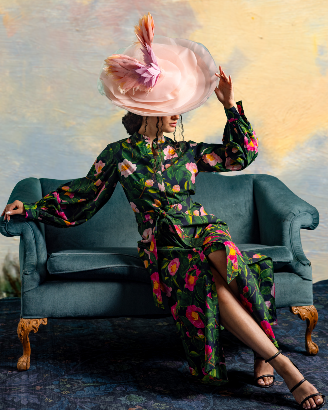 Woman sitting on a vintage blue sofa wearing a dark floral dress and a large, elaborate hat with pink flowers and feathers, posing with one hand resting on the arm of the sofa and the other holding the brim of her hat.