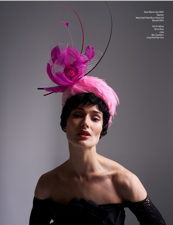 A woman with short dark hair and light skin wearing an elaborate pink fascinator with purple flowers, feathers, and netting, posed against a plain gray background.