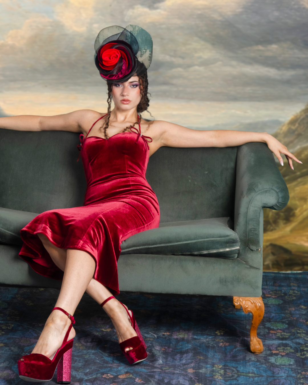 A woman in a red velvet dress and matching high heels sits on a dark green vintage sofa. She wears an elaborate black and red floral hat and has curly hair. The background appears to be a painted landscape scene.