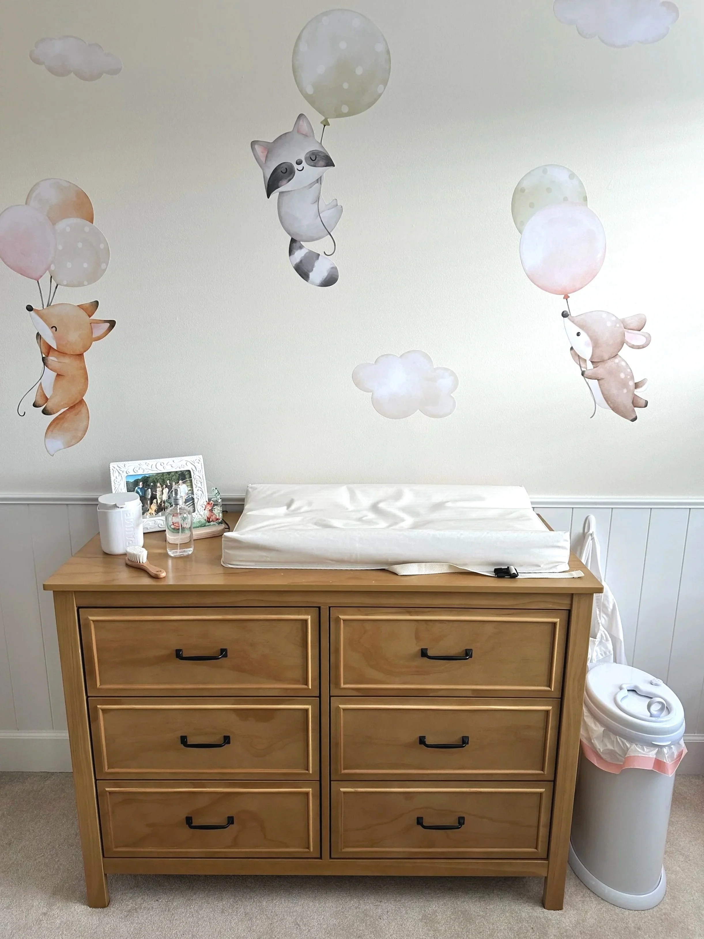 A nursery changing station with a brown dresser, wall decals, a trash can, and changing pad.