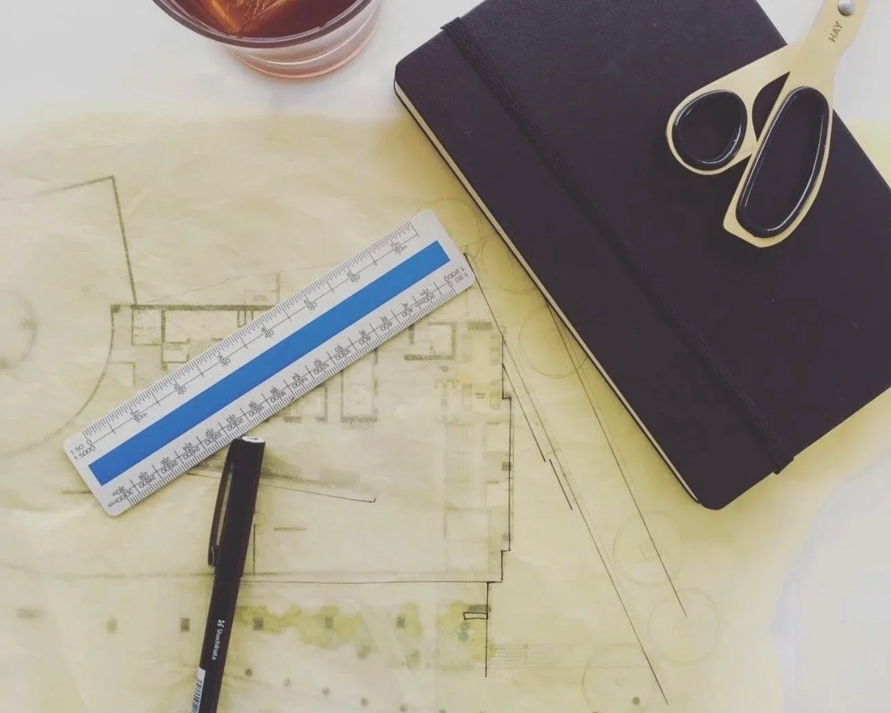Architectural blueprint with a black notebook, scissors, a pen, a scale ruler, and a drink on top.