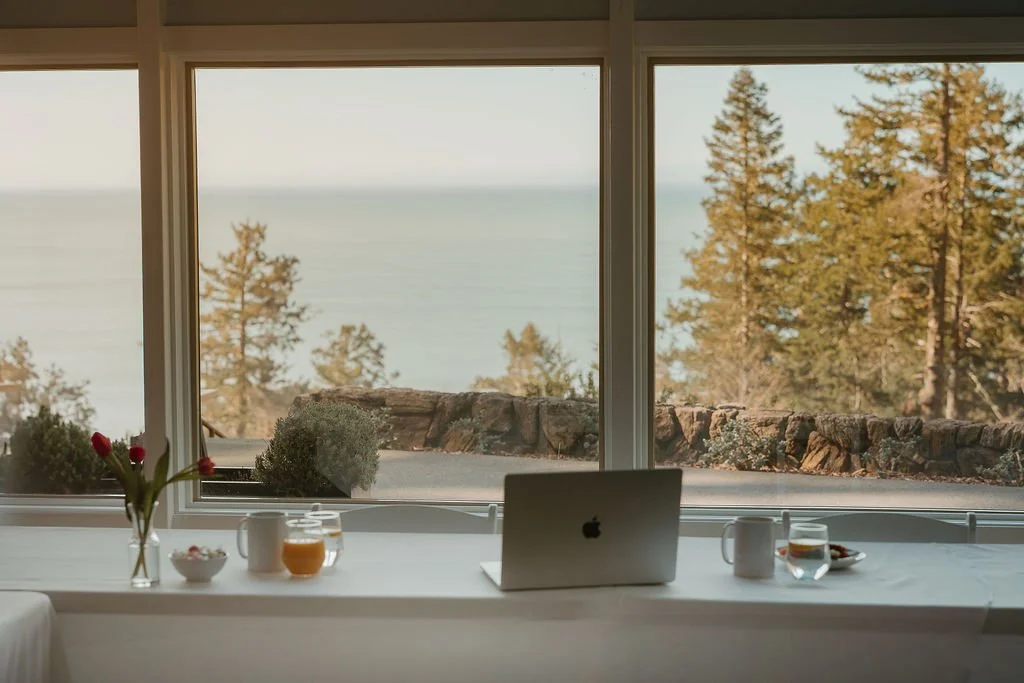 A breakfast table with a laptop, mugs, a bowl, and a vase with flowers, overlooking a scenic view of trees and water through large windows.