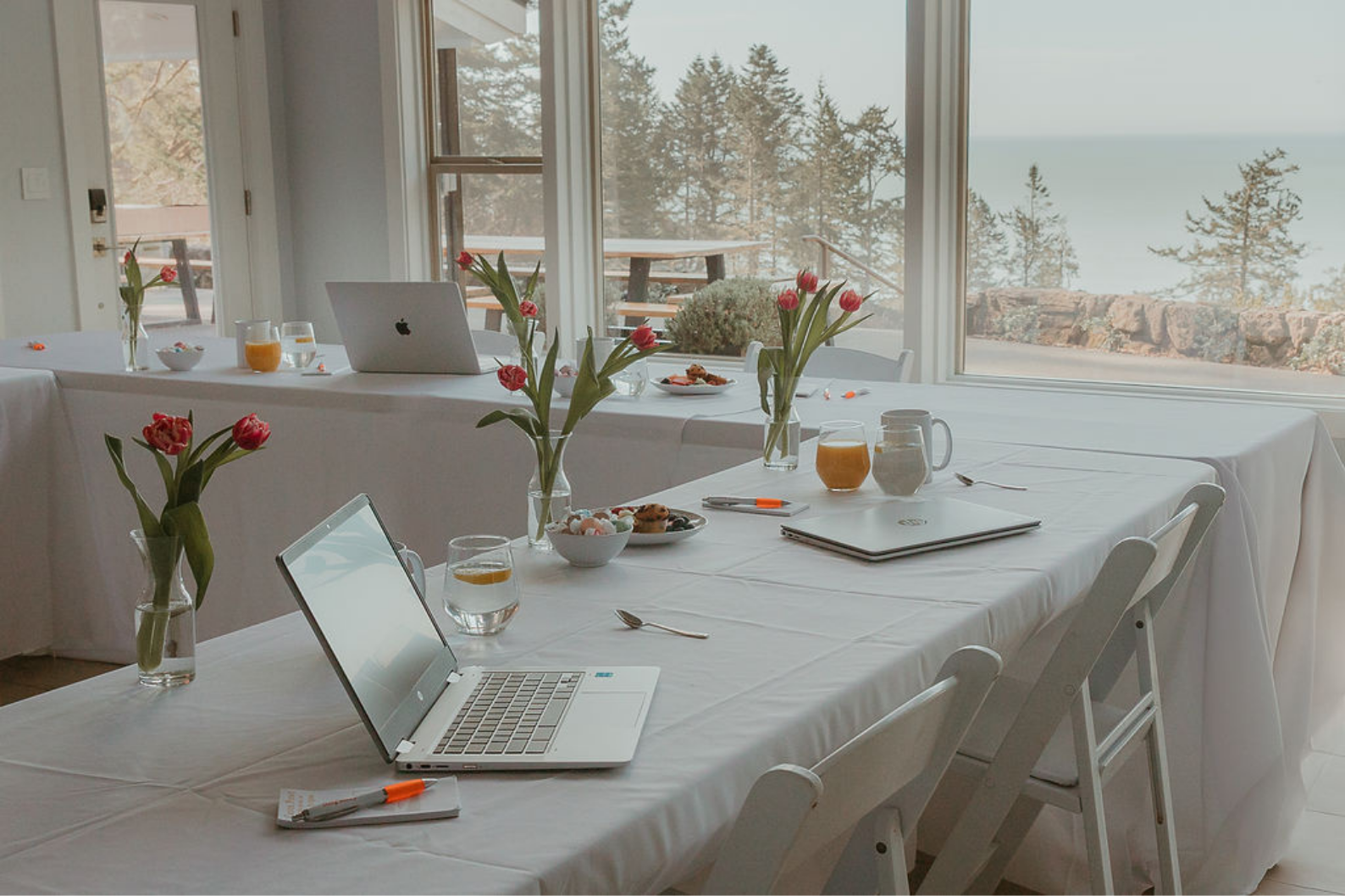 A decorated conference or meeting room with white draped tables, laptops, notebooks, glasses of water and juice, vases of pink tulips, and plates of cookies, with large windows showing trees and a body of water outside.