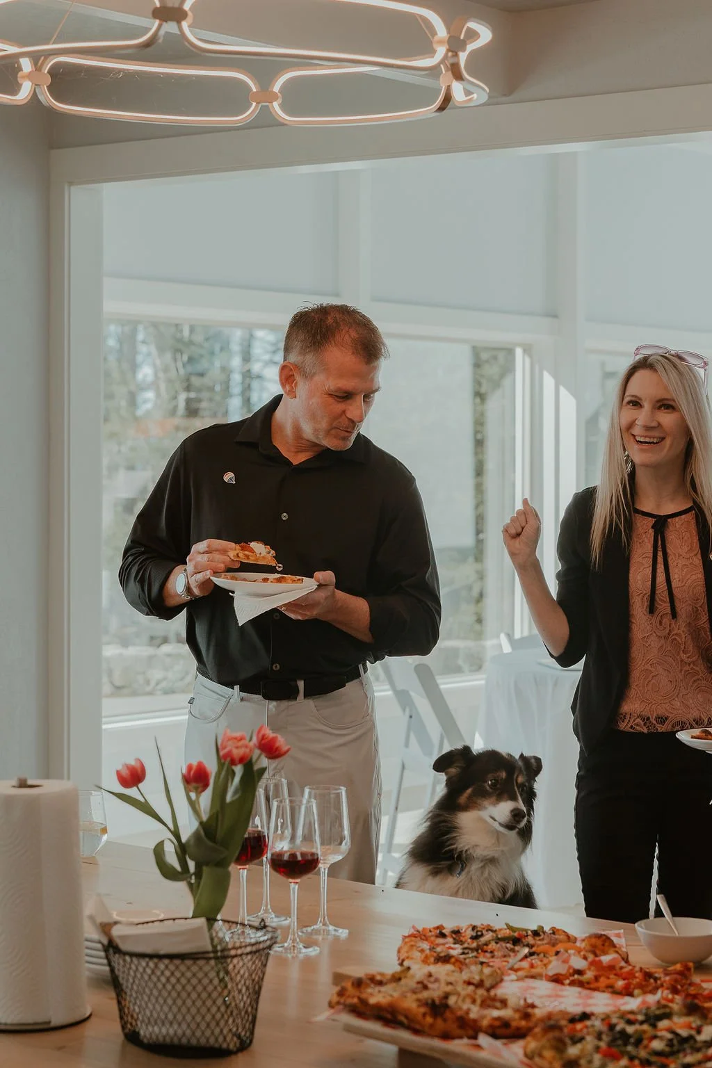 Two people, a man and a woman, standing near a table with pizza, wine glasses, and a vase with pink tulips in a bright room with large windows. The man is holding a plate with food, and the woman is smiling with her fist raised in a celebratory manner. A dog is sitting at the table.