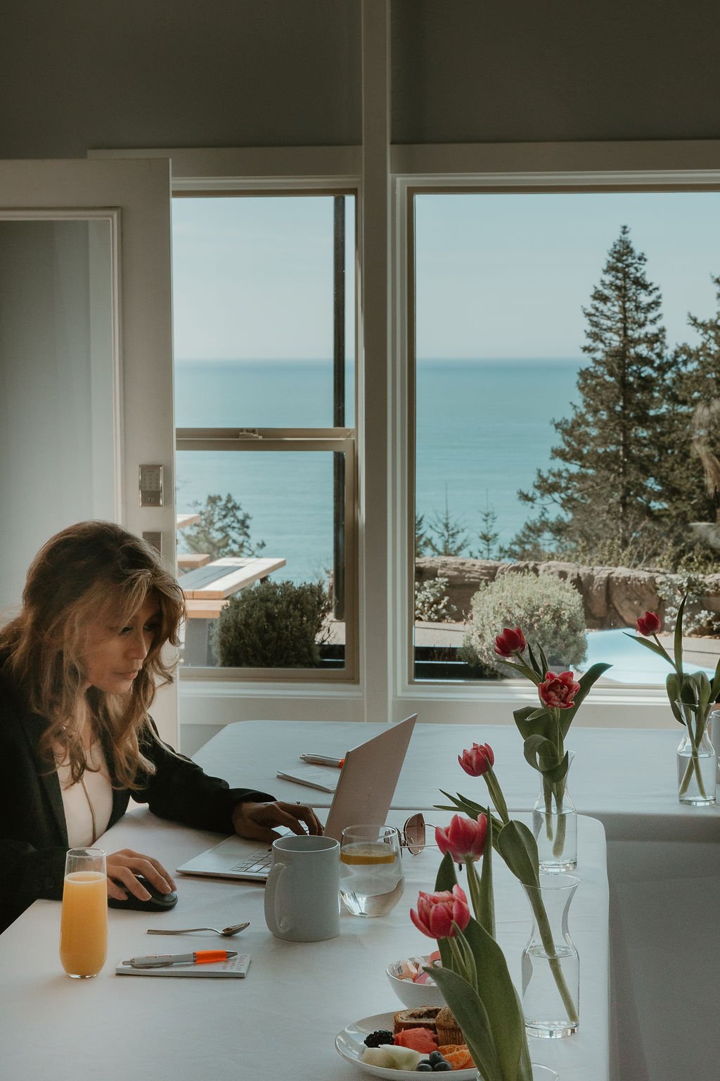 A woman working on a laptop at a dining table with snacks and drinks, with large windows showing a view of trees and the ocean outside.