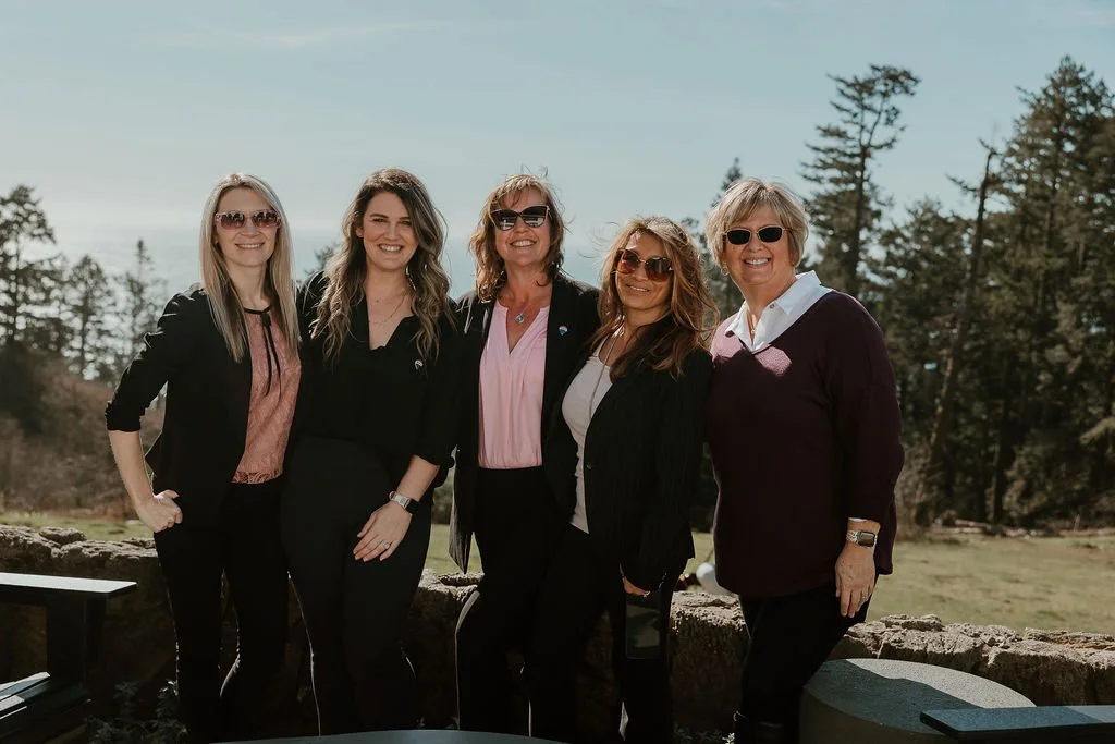 Five women standing outdoors in a line, smiling, with trees in the background, wearing casual and semi-formal attire, sunglasses, and posing during daytime.